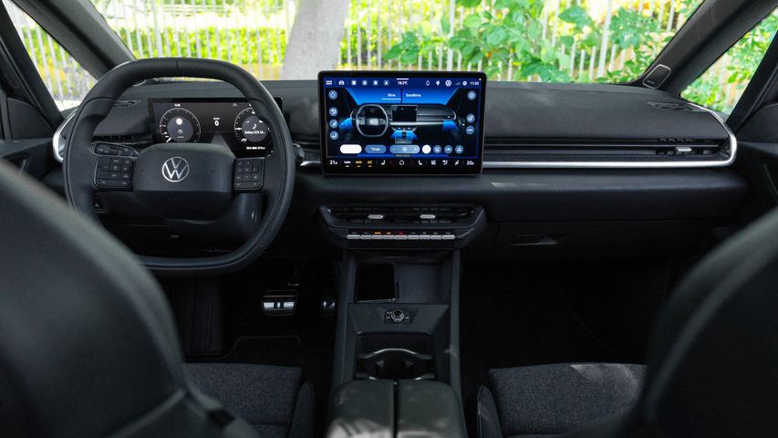 Интериор на модерен автомобил Volkswagen, заснет от задните седалки към таблото. В центъра има голям вертикален сензорен екран с навигация и мултимедия, а пред него – волан с логото на марката. През предното стъкло се вижда зелена растителност и слънчева светлина отвън.