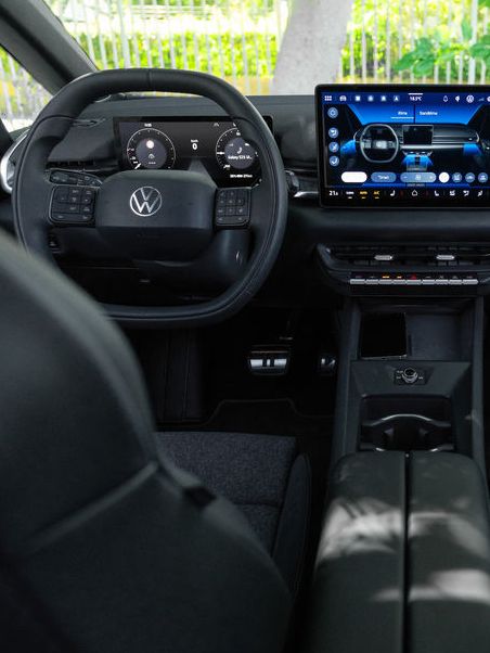 Интериор на модерен автомобил Volkswagen, заснет от задните седалки към таблото. В центъра има голям вертикален сензорен екран с навигация и мултимедия, а пред него – волан с логото на марката. През предното стъкло се вижда зелена растителност и слънчева светлина отвън.