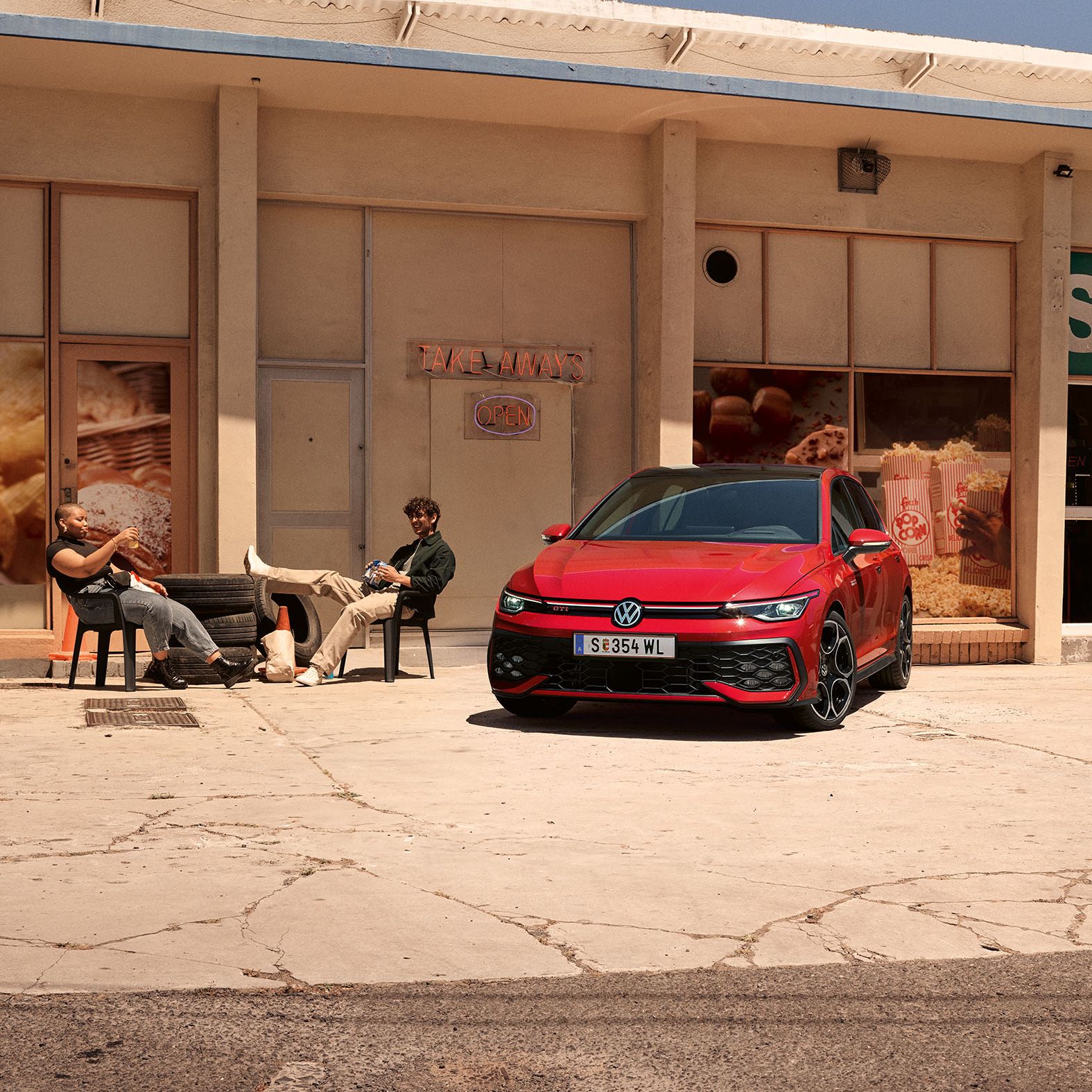 Zwei Personen sitzen auf Plastikstühlen neben einem Golf GTI, dahinter befindet sich ein Shop.