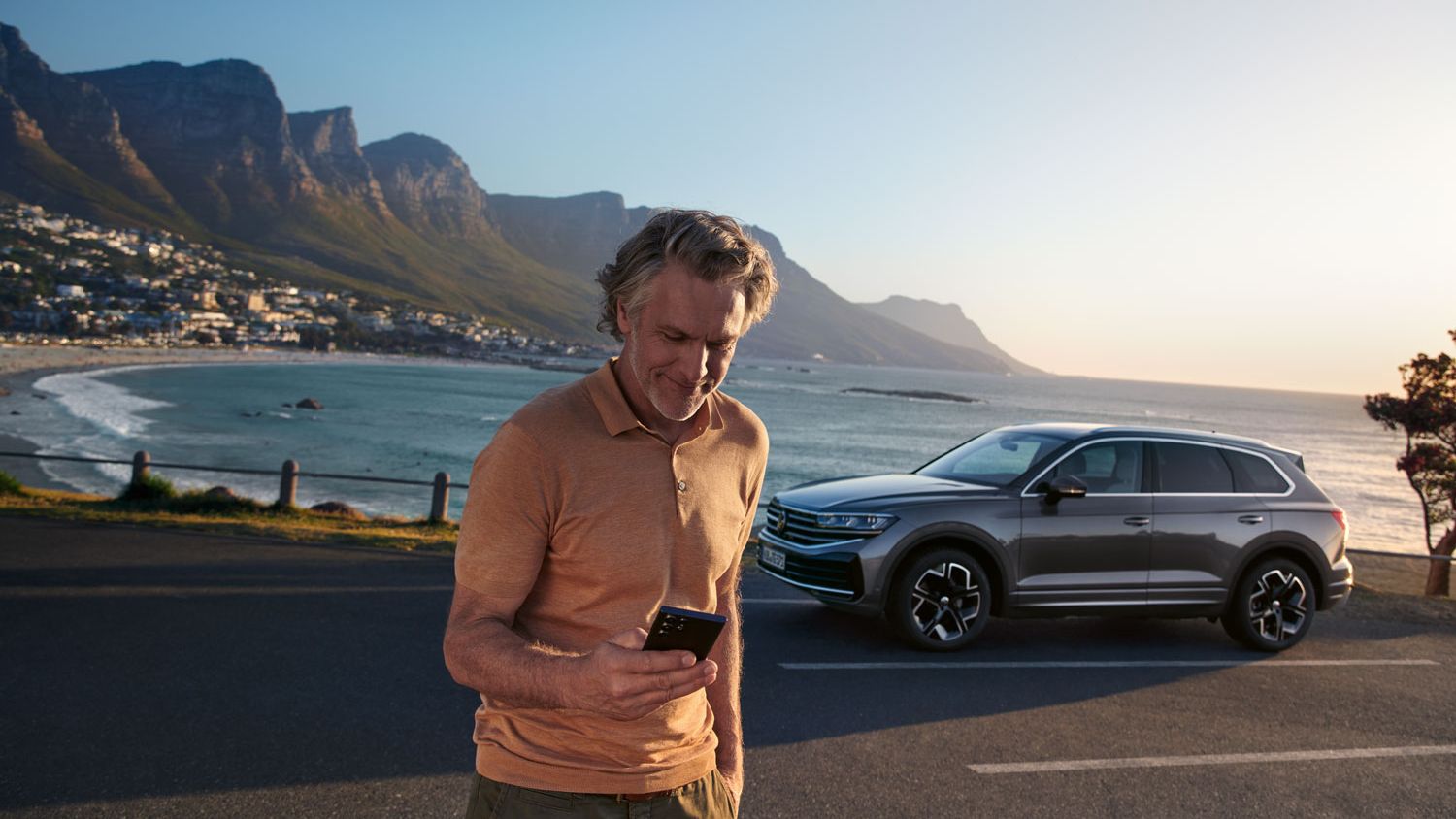Мъж гледа в смартфона си, зад него е VW Touareg Elegance на фона на езеро.