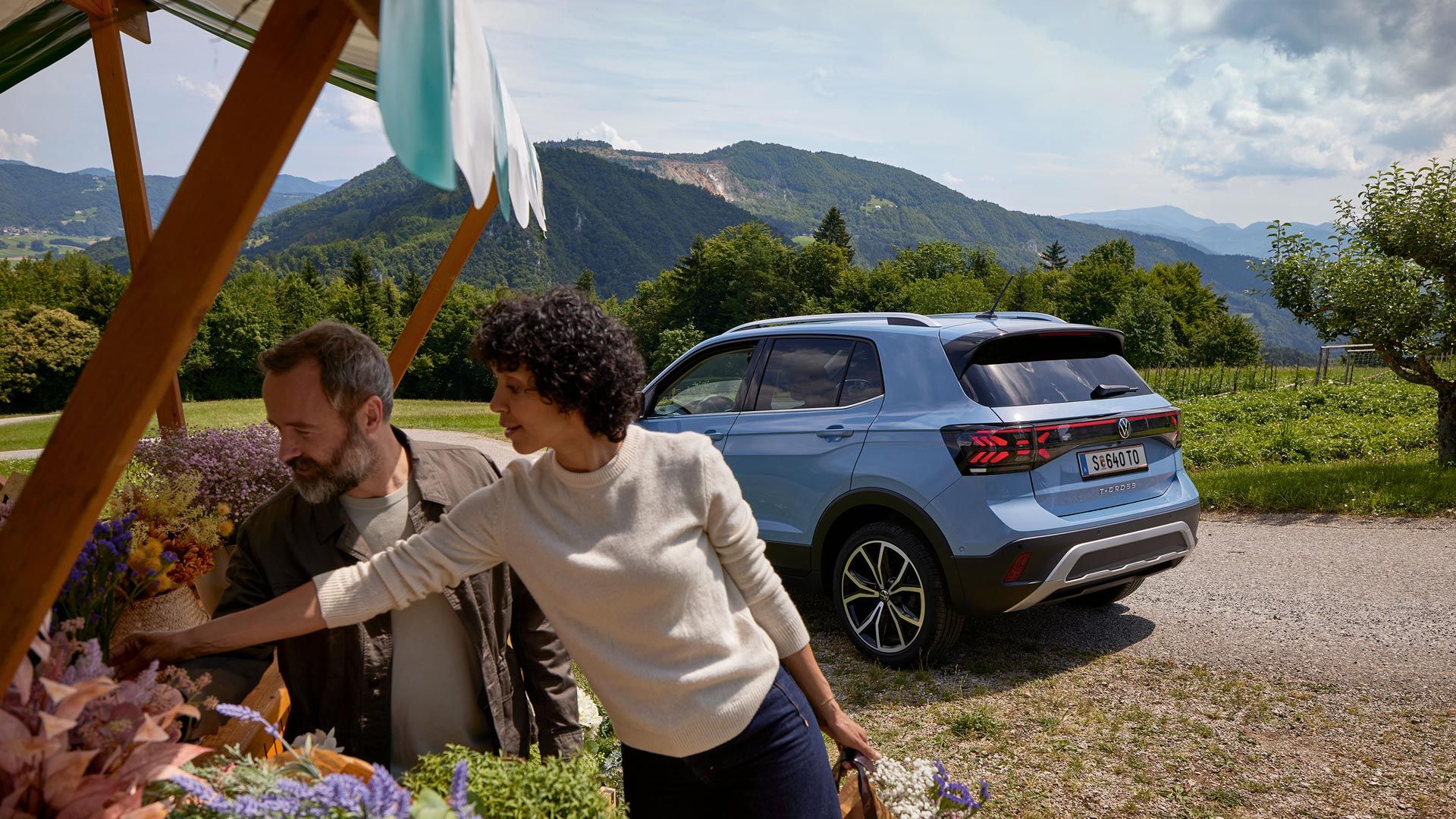 Мъж и жена стоят на щанд за цветя, зад тях е син VW T-Cross на фона на планински пейзаж.