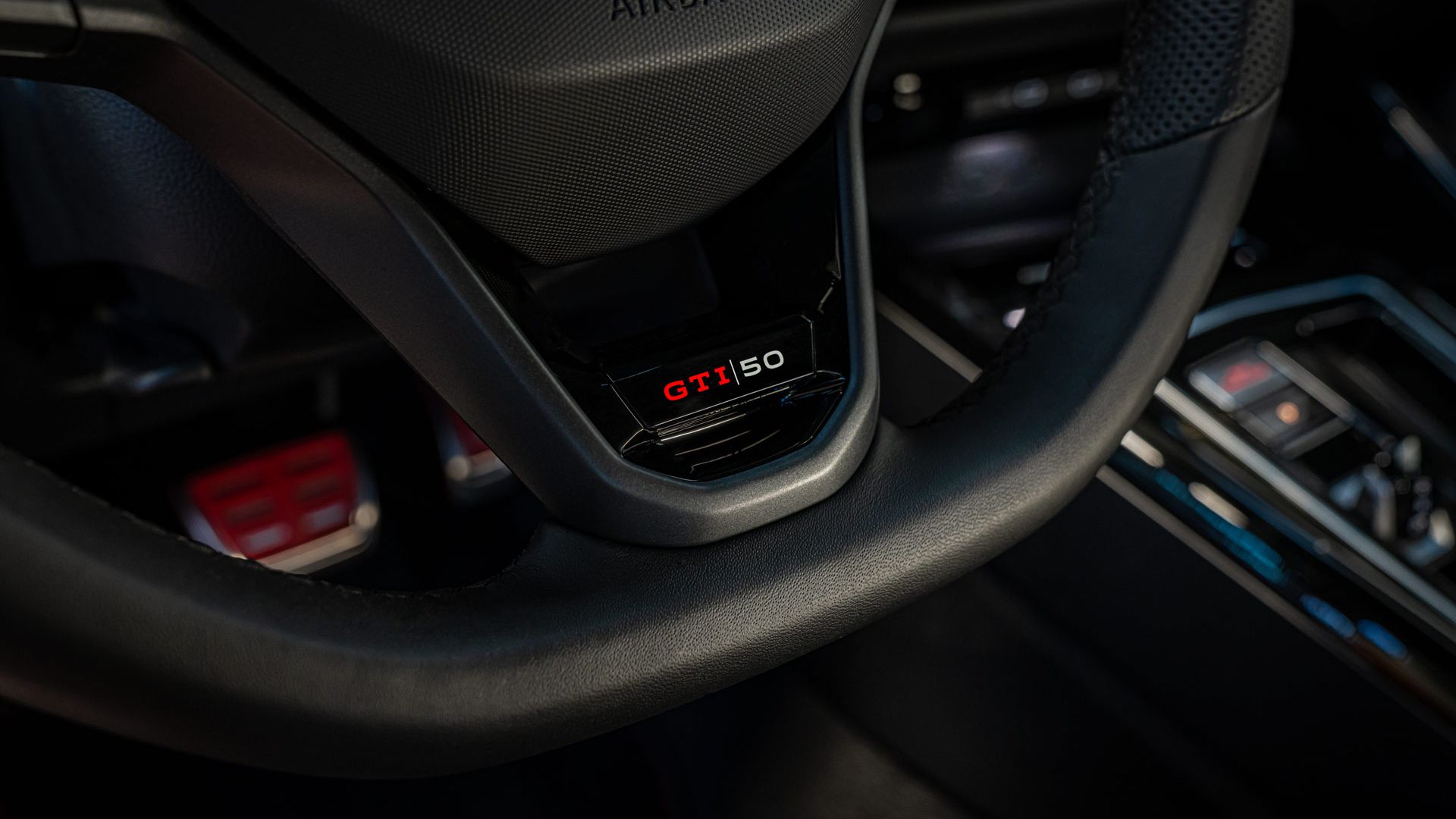 Nahaufnahme eines Lenkrads mit der Aufschrift "GTI 50" in einem Auto. Bedienknöpfe und die dahinterliegende Konsole sind ebenfalls teilweise sichtbar. Der Airbag-Bereich ist mit dem Schriftzug "AIRBAG" markiert.