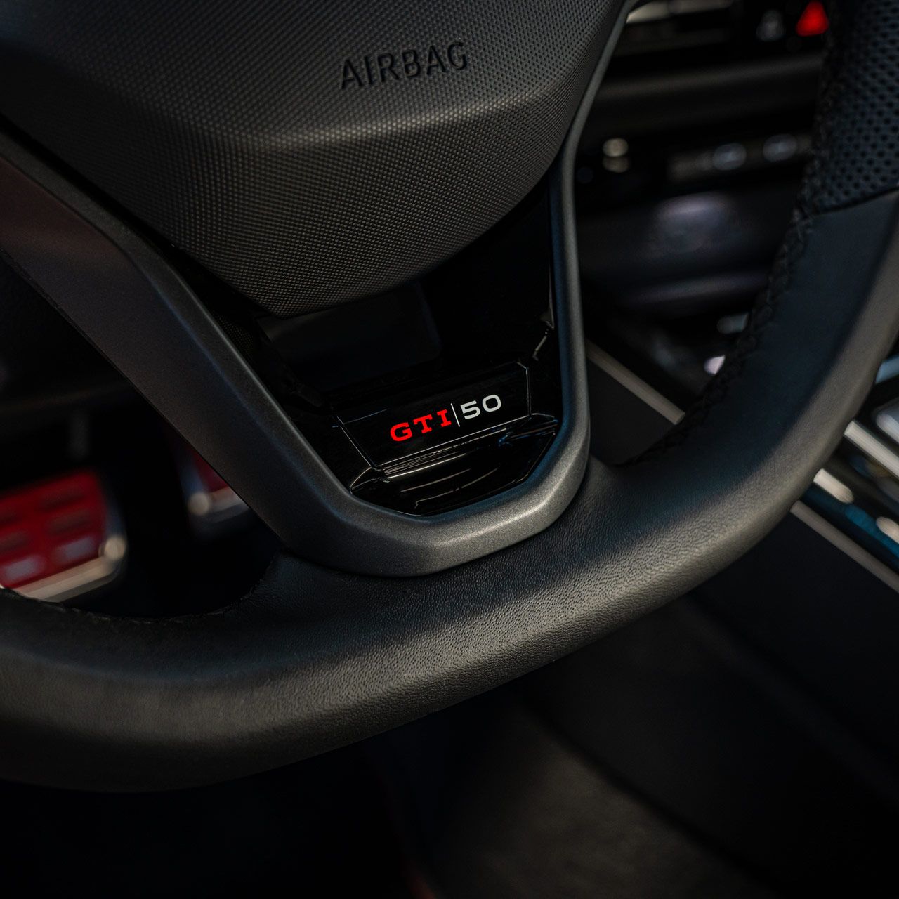 Nahaufnahme eines Lenkrads mit der Aufschrift "GTI 50" in einem Auto. Bedienknöpfe und die dahinterliegende Konsole sind ebenfalls teilweise sichtbar. Der Airbag-Bereich ist mit dem Schriftzug "AIRBAG" markiert.