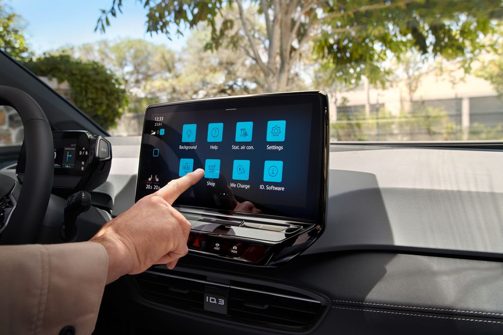 Екран на инфотеймънт системата на автомобил, на който човек докосва иконката за настройка. На дисплея са показани различни иконки, като фона на екрана е син. Отзад се вижда парк с дървета.