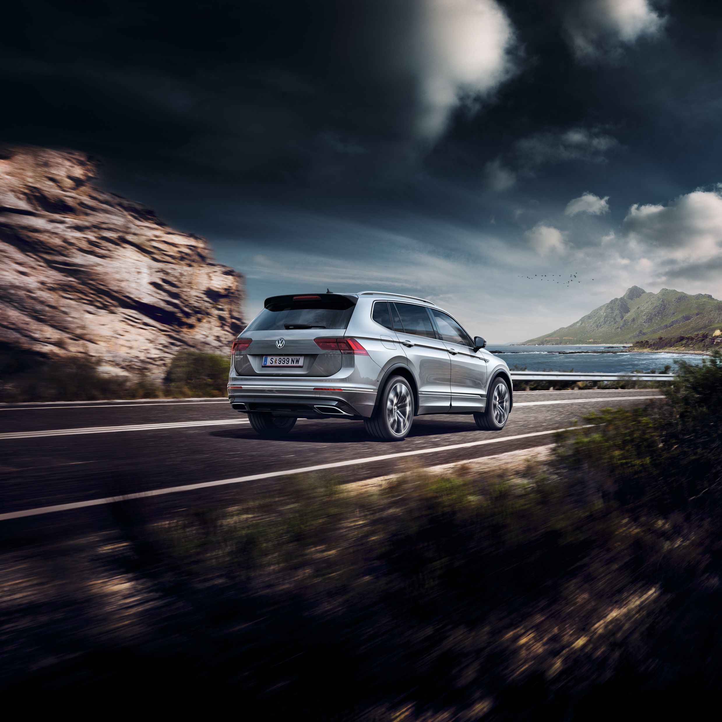 Сребрист Volkswagen Tiguan в движение по живописен път до море и планини.