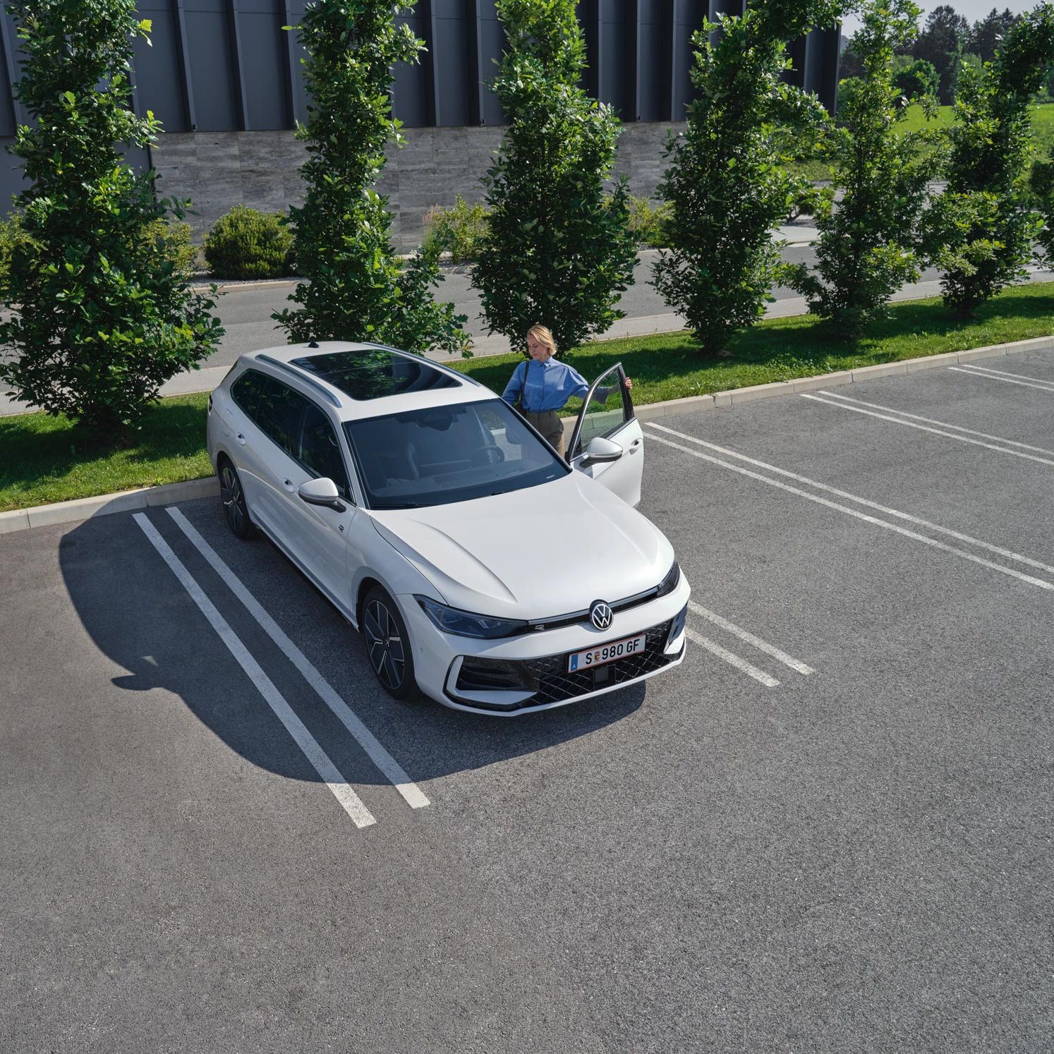 Поглед отгоре към бял VW Passat b9 на паркомясто с жена, качваща се на шофьорското място.