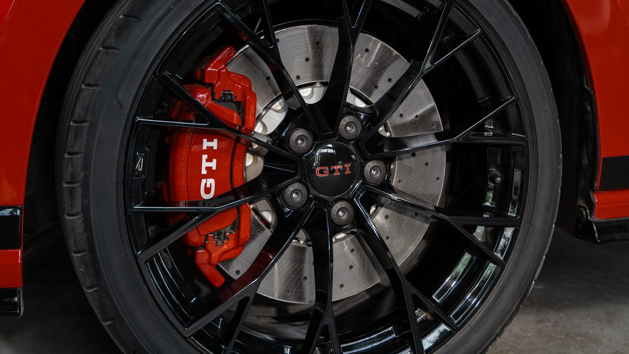 Nahaufnahme eines roten Golf GTI mit schwarzen Rädern. Auf der Bremszange ist das GTI-Logo zu sehen. Das Fahrzeug steht in einer industriellen Umgebung.