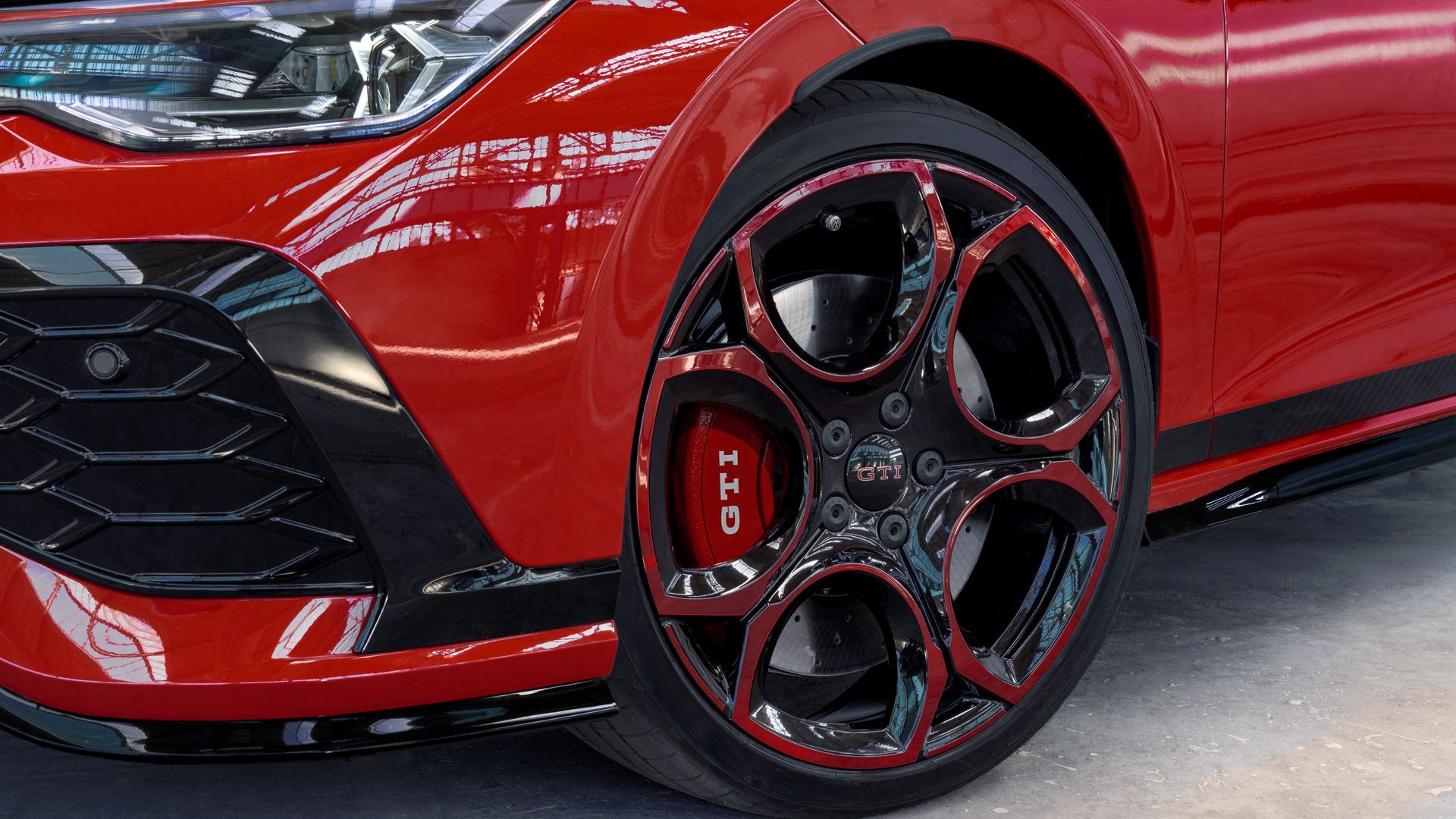 Nahaufnahme eines roten Golf GTI, das eine markante schwarze und rote Felge mit dem GTI-Logo zeigt. Die Frontschürze ist teils sichtbar und hat eine Wabenstruktur. Das Auto steht in einer hellen Umgebung mit viel natürlichem Licht.