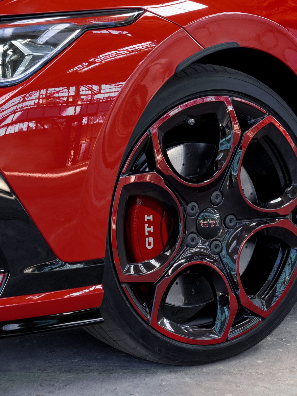 Nahaufnahme eines roten Golf GTI, das eine markante schwarze und rote Felge mit dem GTI-Logo zeigt. Die Frontschürze ist teils sichtbar und hat eine Wabenstruktur. Das Auto steht in einer hellen Umgebung mit viel natürlichem Licht.