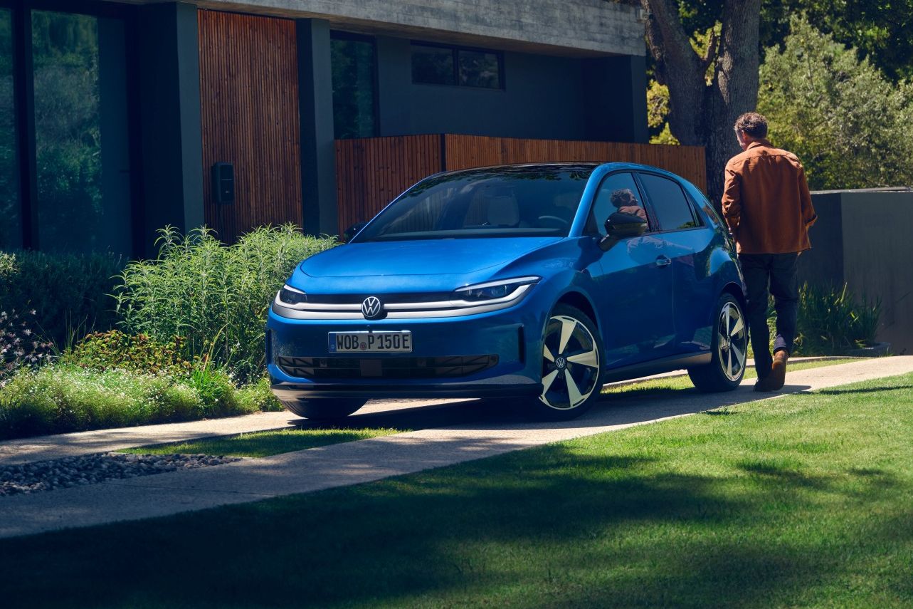 Син Volkswagen ID.Polo е паркиран на алея пред модерна къща с големи стъклени прозорци и зелена растителност по покрива. Мъж, облечен в кафяво яке, стои до колата и гледа към къщата. Наоколо има добре поддържана морава, дървета и планини на заден план.