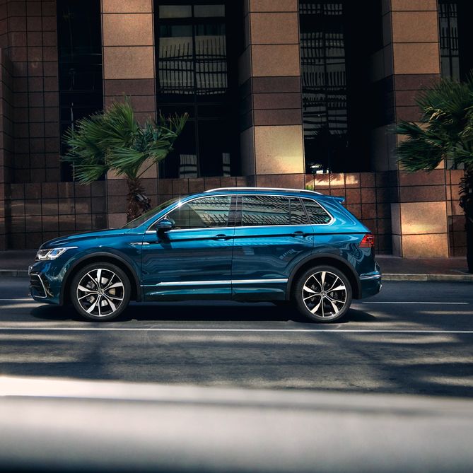 Син Volkswagen Tiguan R, заснет в профил на празна улица пред модерна офис сграда.