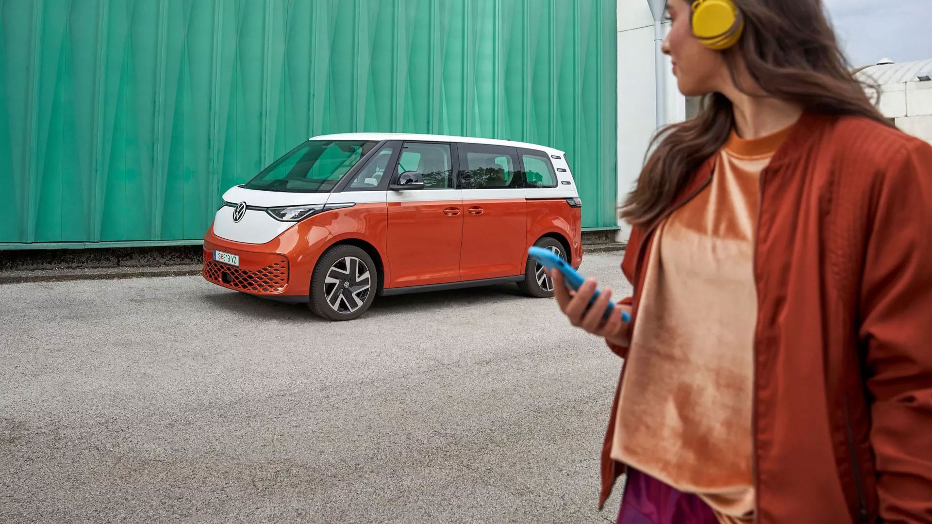 VW ID. Buzz в бяло и оранжева, жена гледа към автомобила 