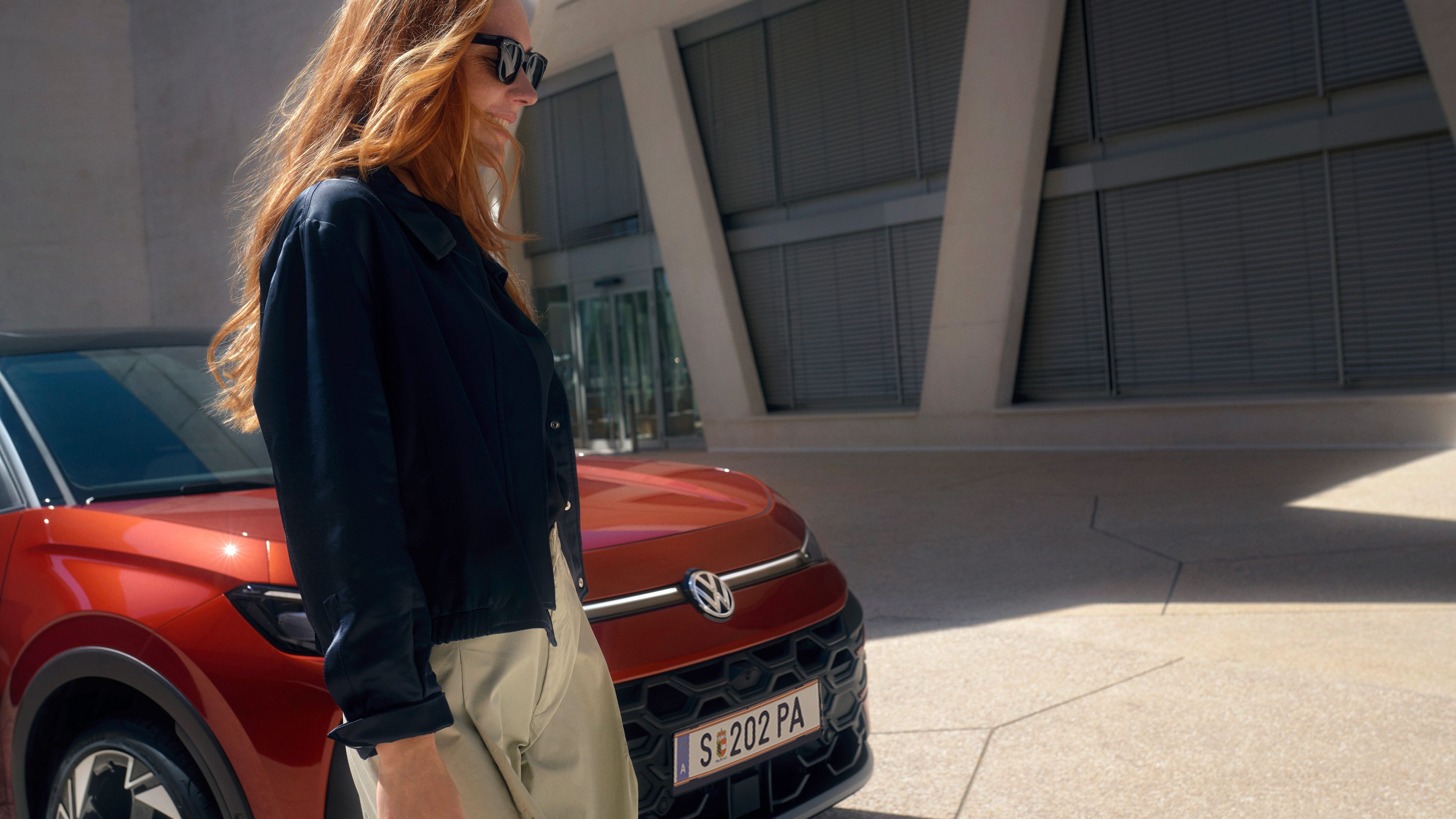 Жена със слънчеви очила до червен Volkswagen T-Roc пред модерна сграда.