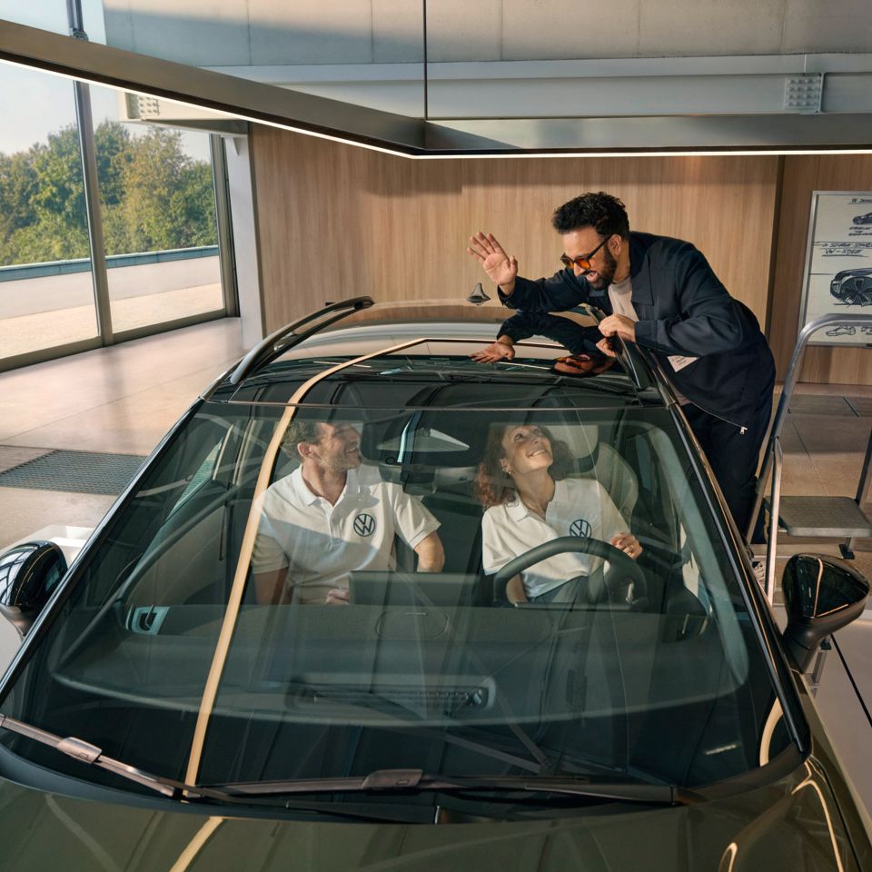Мъж стои на стълба до VW ID. Cross и маха през панорамния покрив на хората, които седят вътре в автомобила.