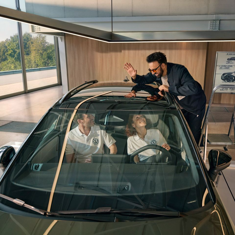 Мъж стои на стълба до VW ID. Cross и маха през панорамния покрив на хората, които седят вътре в автомобила.