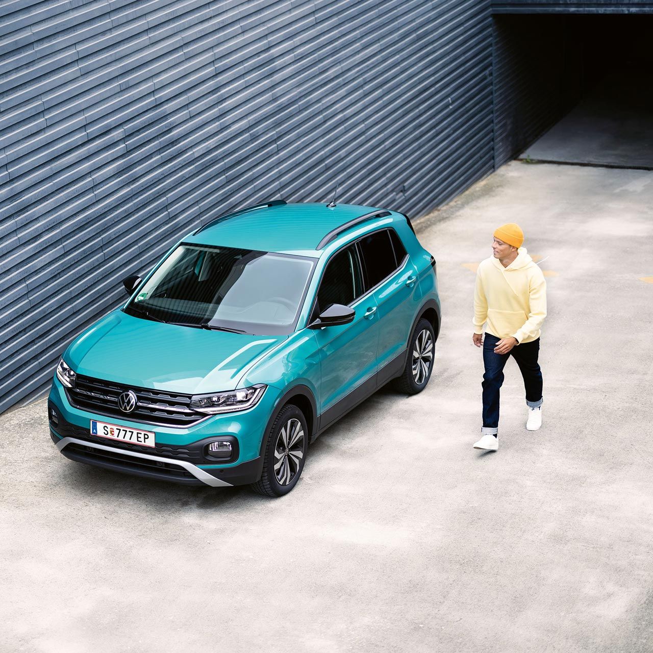 Зелен Volkswagen T-Cross, паркиран до индустриална стена, до която върви мъж с жълта шапка.