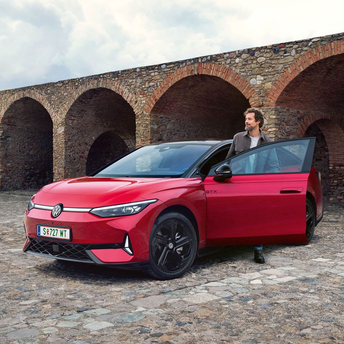 Червен Volkswagen ID.7 GTX паркиран пред каменна арка с водач до отворената врата