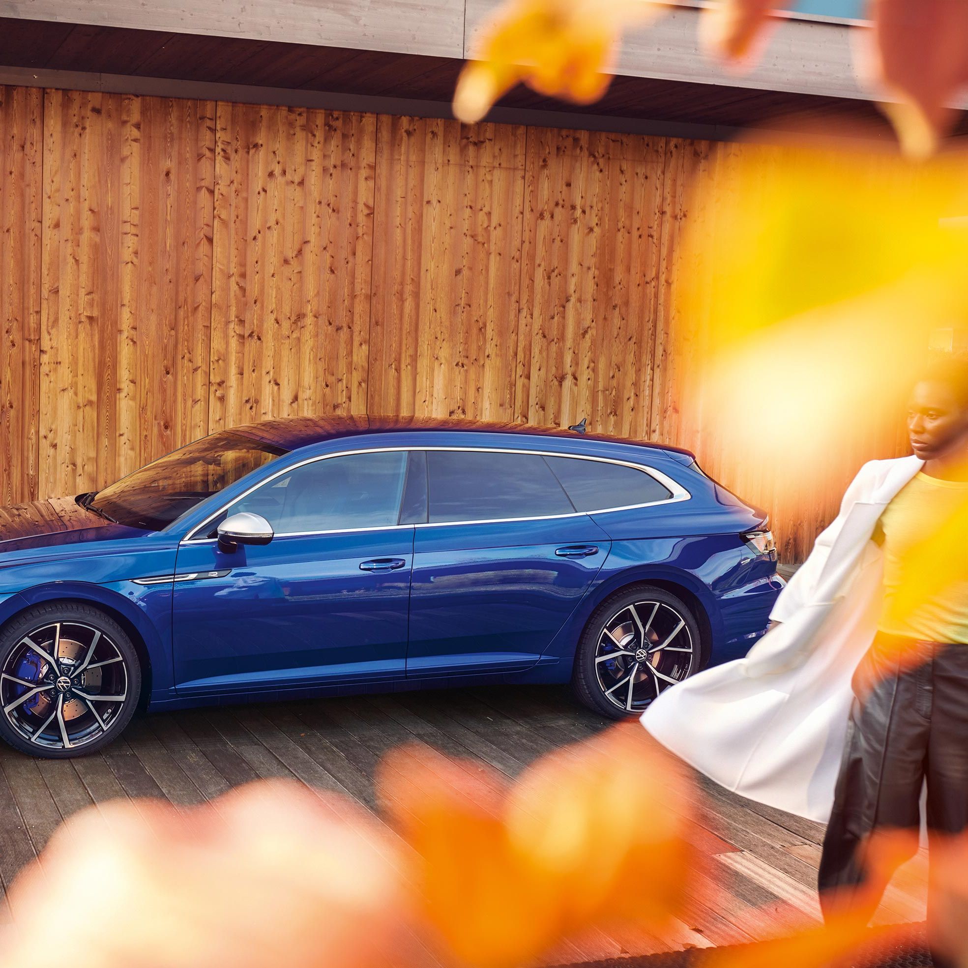 Елегантен син Volkswagen Arteon Shooting Brake паркиран пред модерна дървена сграда с есенни акценти