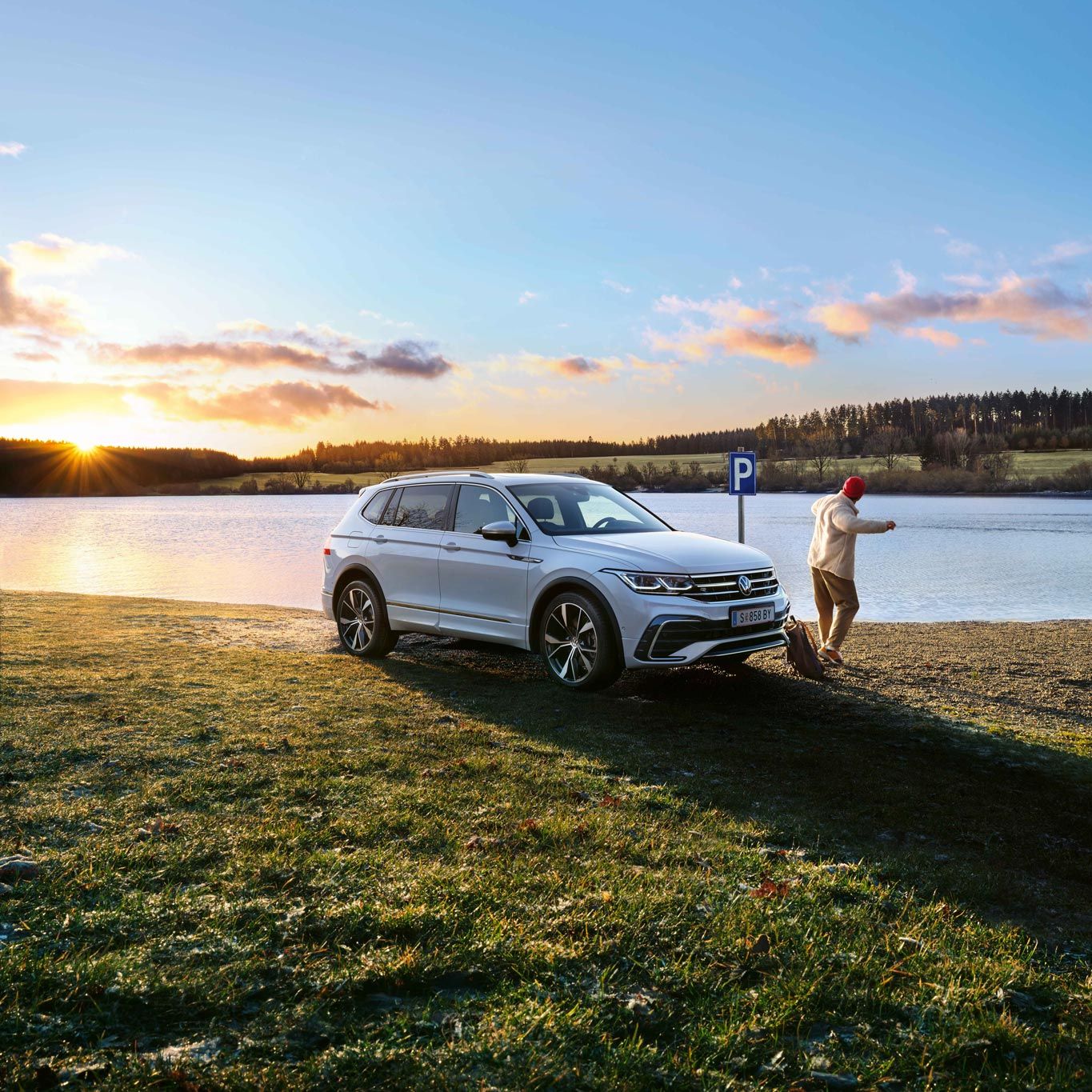 Бял Volkswagen Tiguan паркиран край езеро по залез с човек, който се разхожда наблизо.