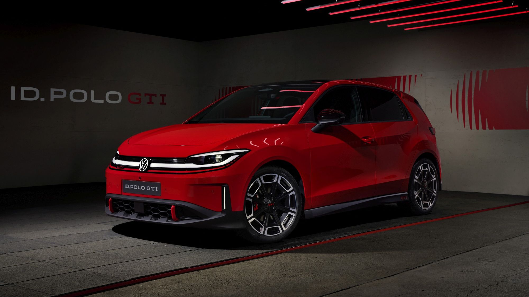 Яркочервен Volkswagen ID. Polo GTI е паркиран в тъмен, модерно осветен шоурум с червени неонови линии по тавана. Колата има агресивен спортен дизайн с LED светлини и големи алуминиеви джанти. На стената зад нея се вижда надпис „ID. POLO GTI“.