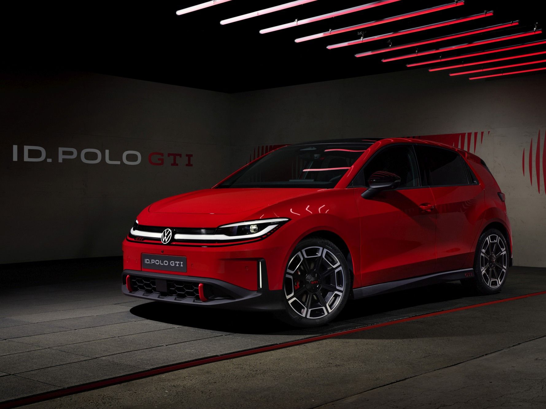 Яркочервен Volkswagen ID. Polo GTI е паркиран в тъмен, модерно осветен шоурум с червени неонови линии по тавана. Колата има агресивен спортен дизайн с LED светлини и големи алуминиеви джанти. На стената зад нея се вижда надпис „ID. POLO GTI“.