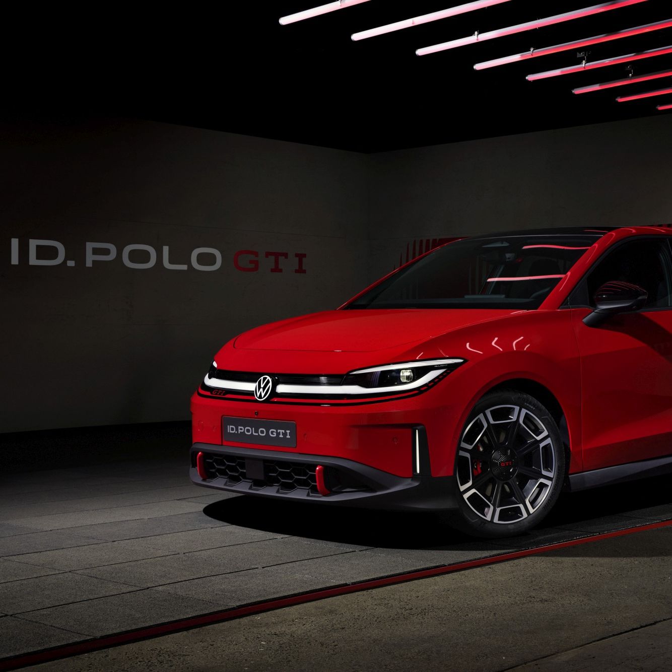 Яркочервен Volkswagen ID. Polo GTI е паркиран в тъмен, модерно осветен шоурум с червени неонови линии по тавана. Колата има агресивен спортен дизайн с LED светлини и големи алуминиеви джанти. На стената зад нея се вижда надпис „ID. POLO GTI“.