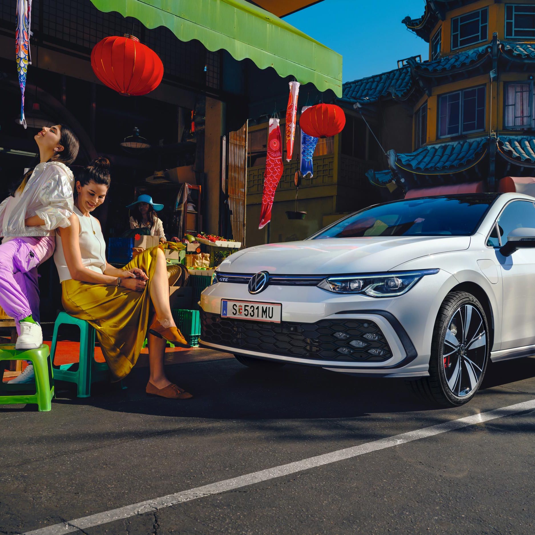 Бял Volkswagen Golf GTE паркиран пред пазар с хора и цветни декорации в оживена градска среда