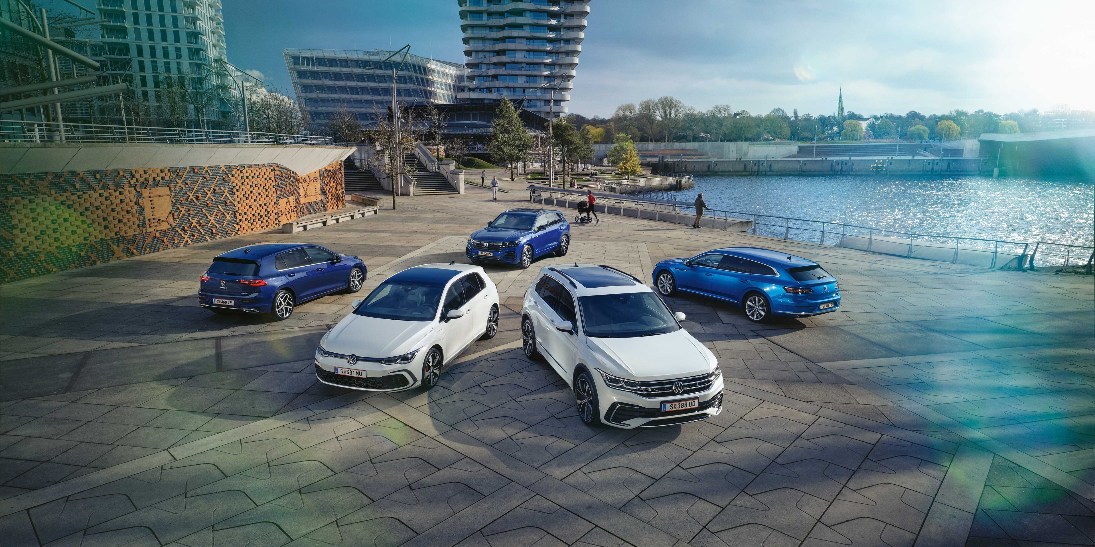 Blick von oben auf drei blaue und zwei weiße VW Hybridmodelle an einer sonnigen Uferpromenade.