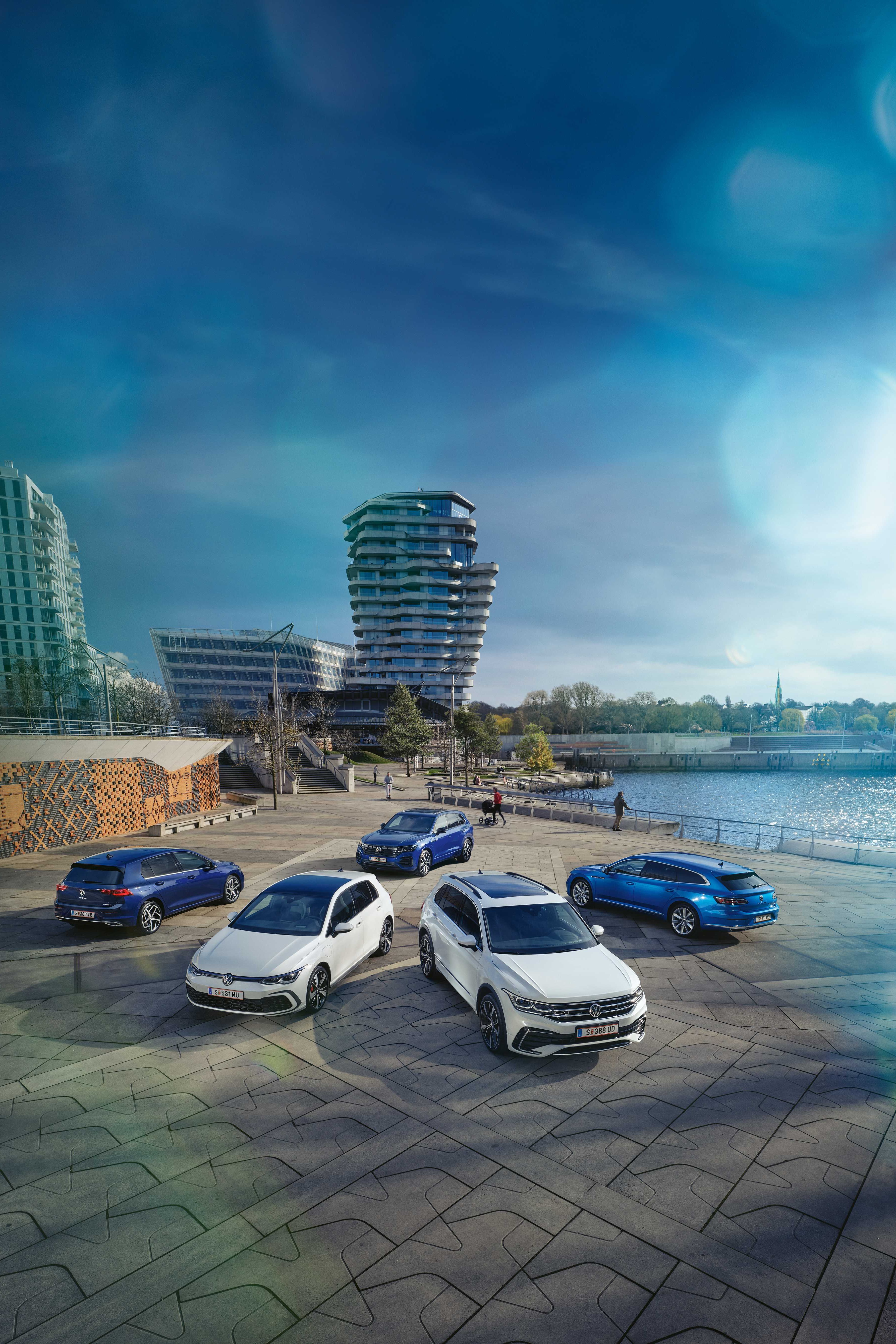 Blick von oben auf drei blaue und zwei weiße VW Hybridmodelle an einer sonnigen Uferpromenade.