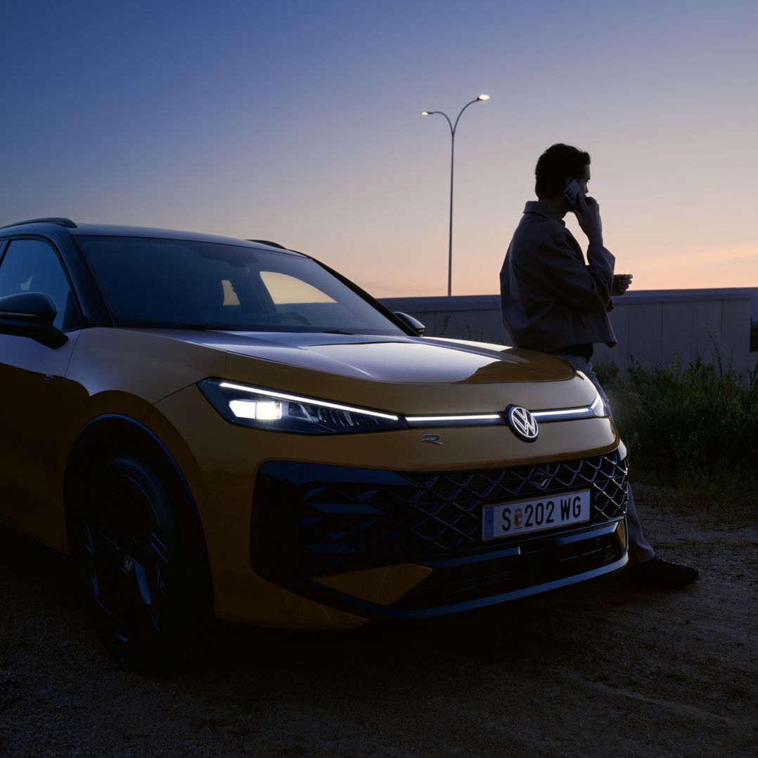 Жълт Volkswagen T-Roc R-Line в здрач с включени предни светлини и силует на мъж до автомобила.