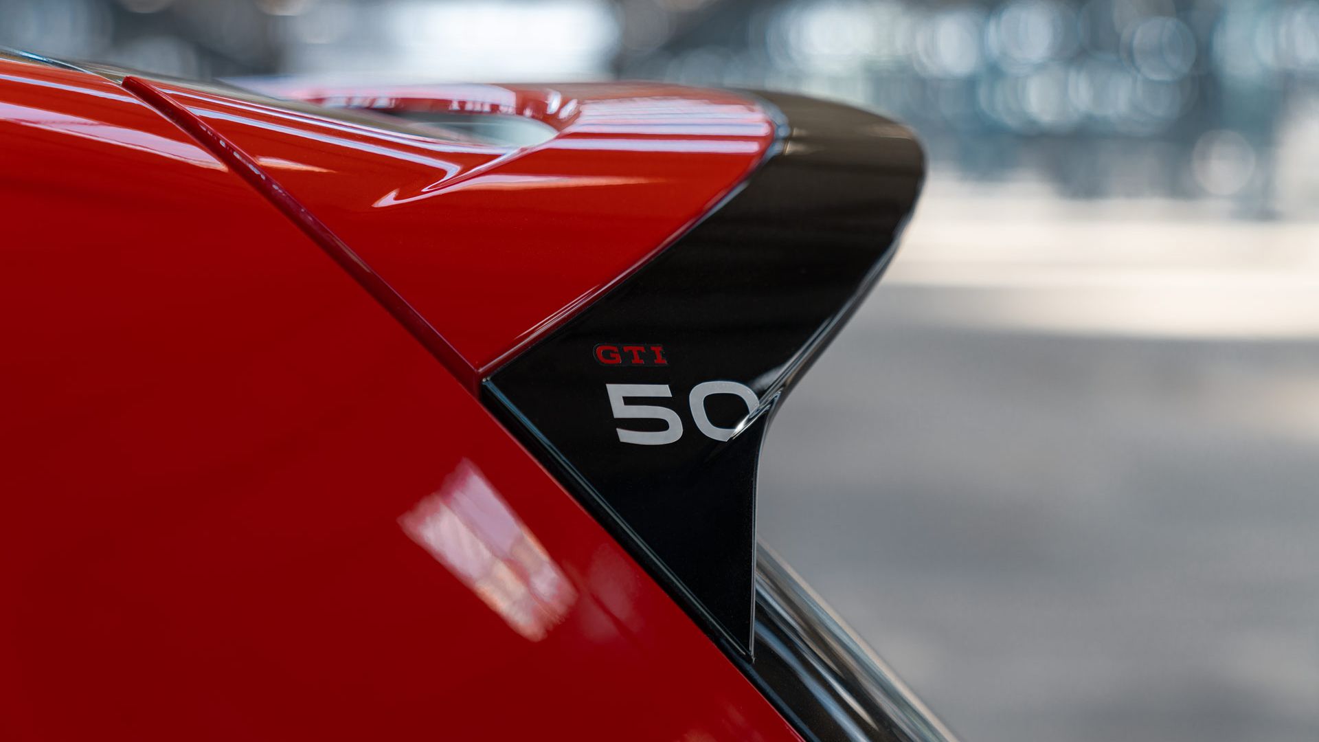 Nahaufnahme eines Golf GTI mit rotem Lack, das Emblem "GTI 50" ist sichtbar. Es scheint sich um einen Spoiler oder eine andere auffällige Karosserieteil zu handeln. Der Hintergrund ist unscharf und bietet wenig visuelle Informationen.