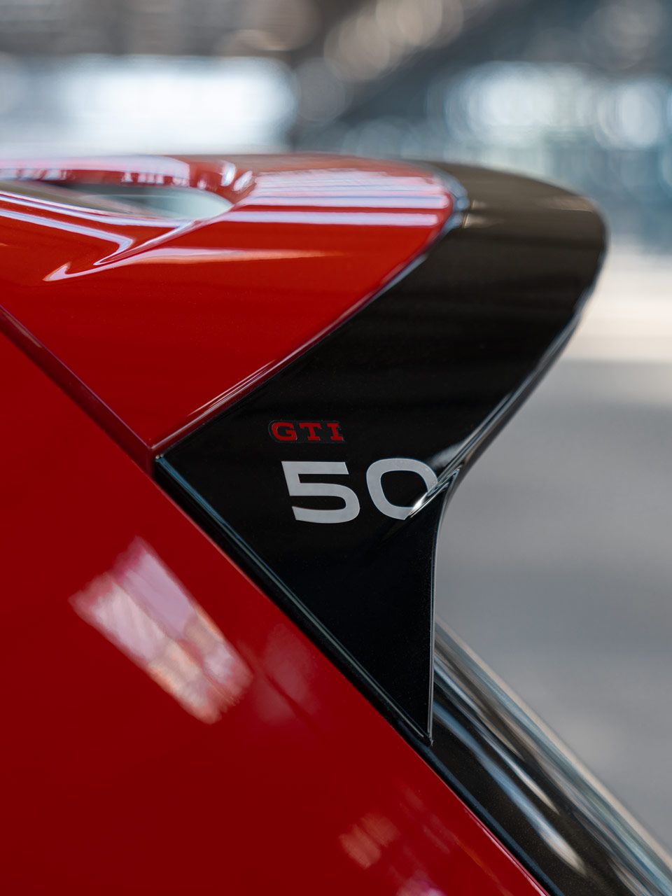Nahaufnahme eines Golf GTI mit rotem Lack, das Emblem "GTI 50" ist sichtbar. Es scheint sich um einen Spoiler oder eine andere auffällige Karosserieteil zu handeln. Der Hintergrund ist unscharf und bietet wenig visuelle Informationen.