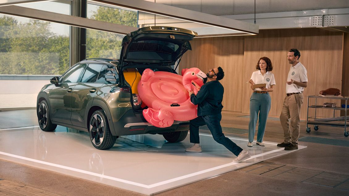Мъж бута надуваемо фламинго в отворения багажник на VW ID. Кръст. Двама души стоят до него и го наблюдават с усмивка.