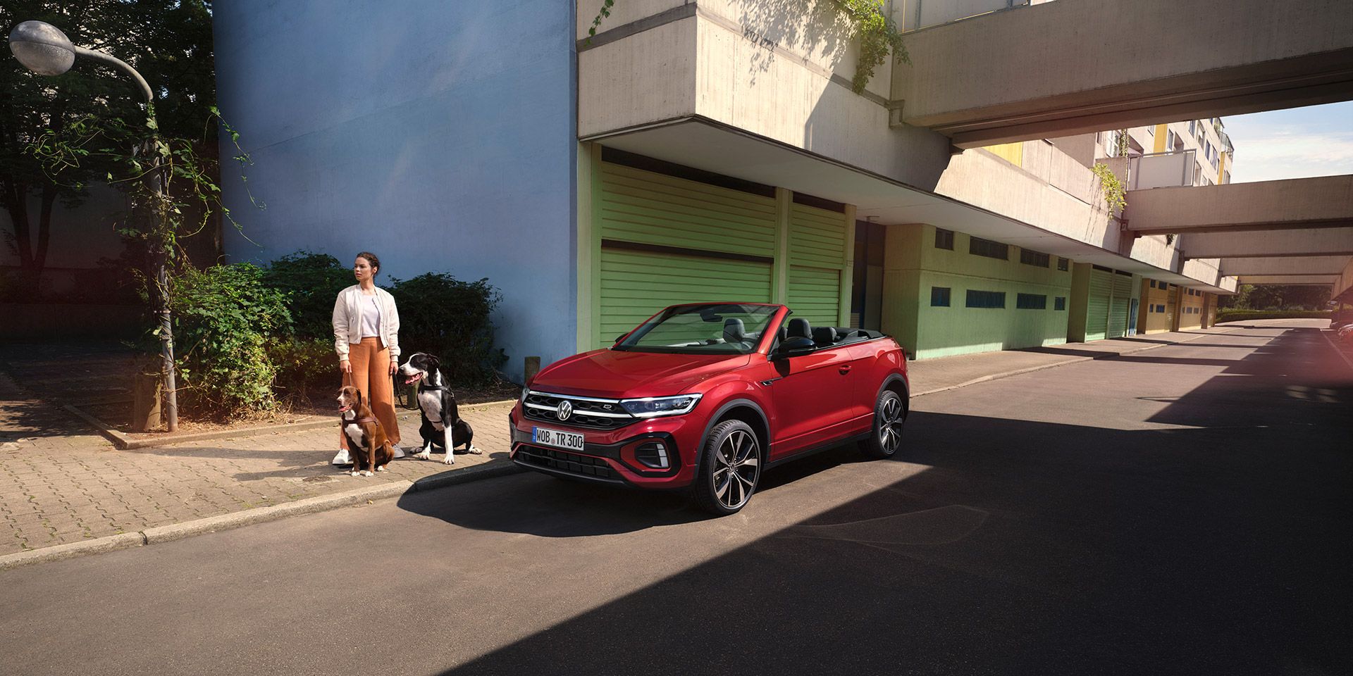 Rotes VW T-Roc Cabriolet mit geöffnetem Dach parkt am Straßenrand. Eine Frau mit zwei Hunden steht daneben. 
