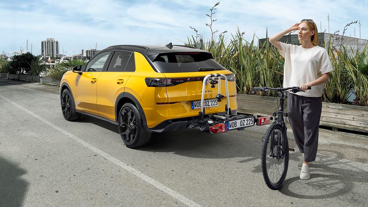 Млада жена с велосипед стои до жълт T-Roc, паркиран на път с прикрепен багажник за велосипеди отзад. Тя гледа в далечината, като си засенчва очите с ръка, а на заден план има зелена растителност и градски силует. Сцената създава усещане за пътуване и активен начин на живот.