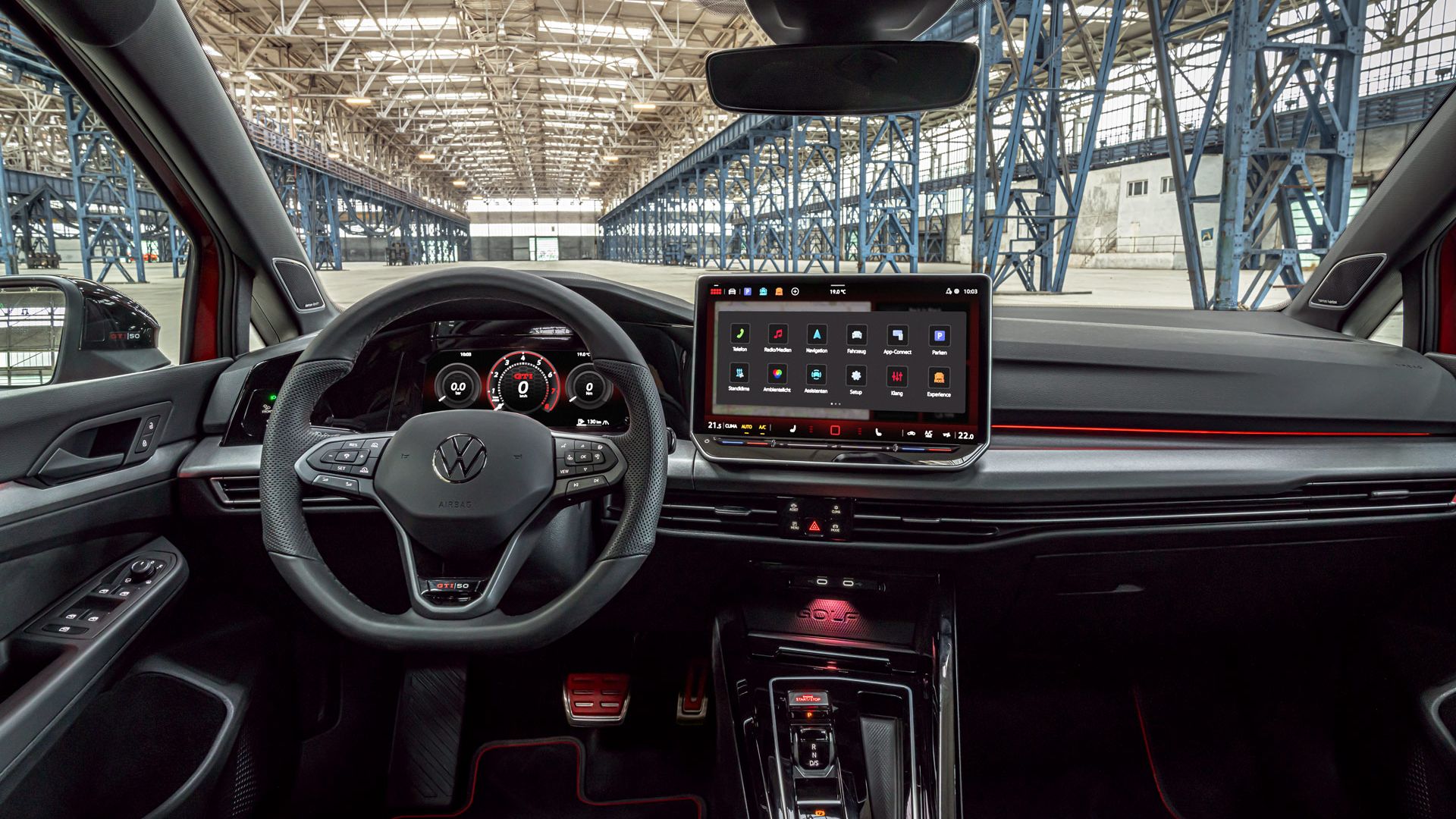 Das Bild zeigt das Cockpit eines modernen Golf GTI. Auffällig ist das große Touchscreen-Bedienfeld und die digitale Instrumententafel. Im Hintergrund ist eine große Industriehalle zu sehen.