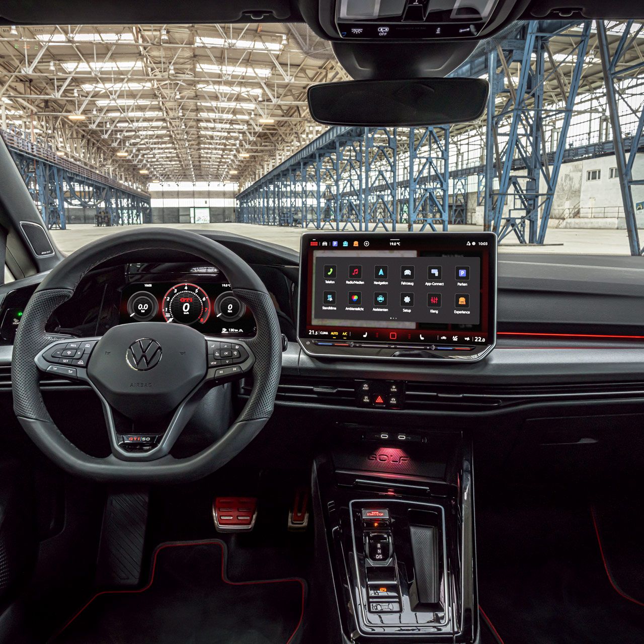 Das Bild zeigt das Cockpit eines modernen Golf GTI. Auffällig ist das große Touchscreen-Bedienfeld und die digitale Instrumententafel. Im Hintergrund ist eine große Industriehalle zu sehen.
