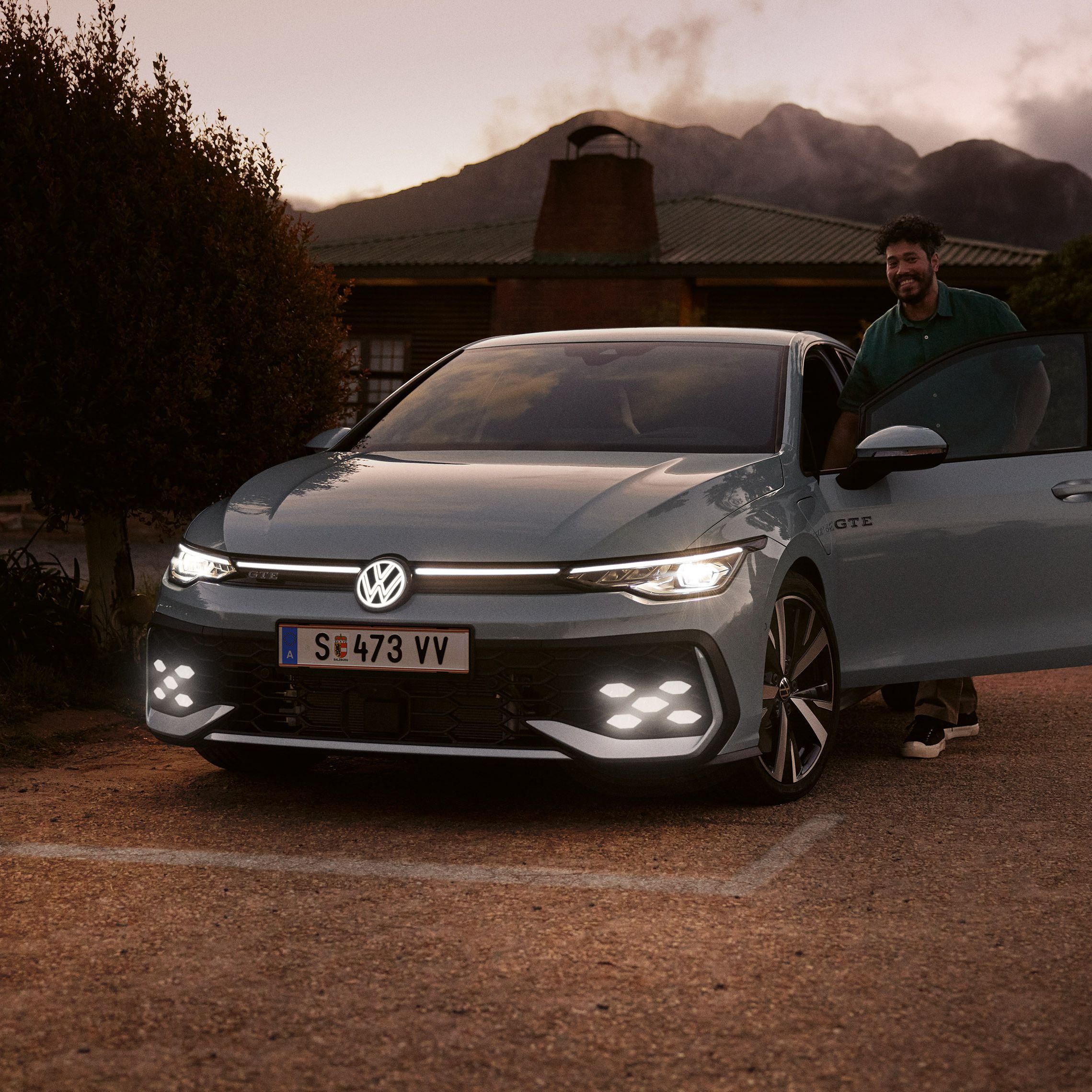 Син Volkswagen Golf GTE с отворена врата и усмихнат водач на фона на планини по здрач