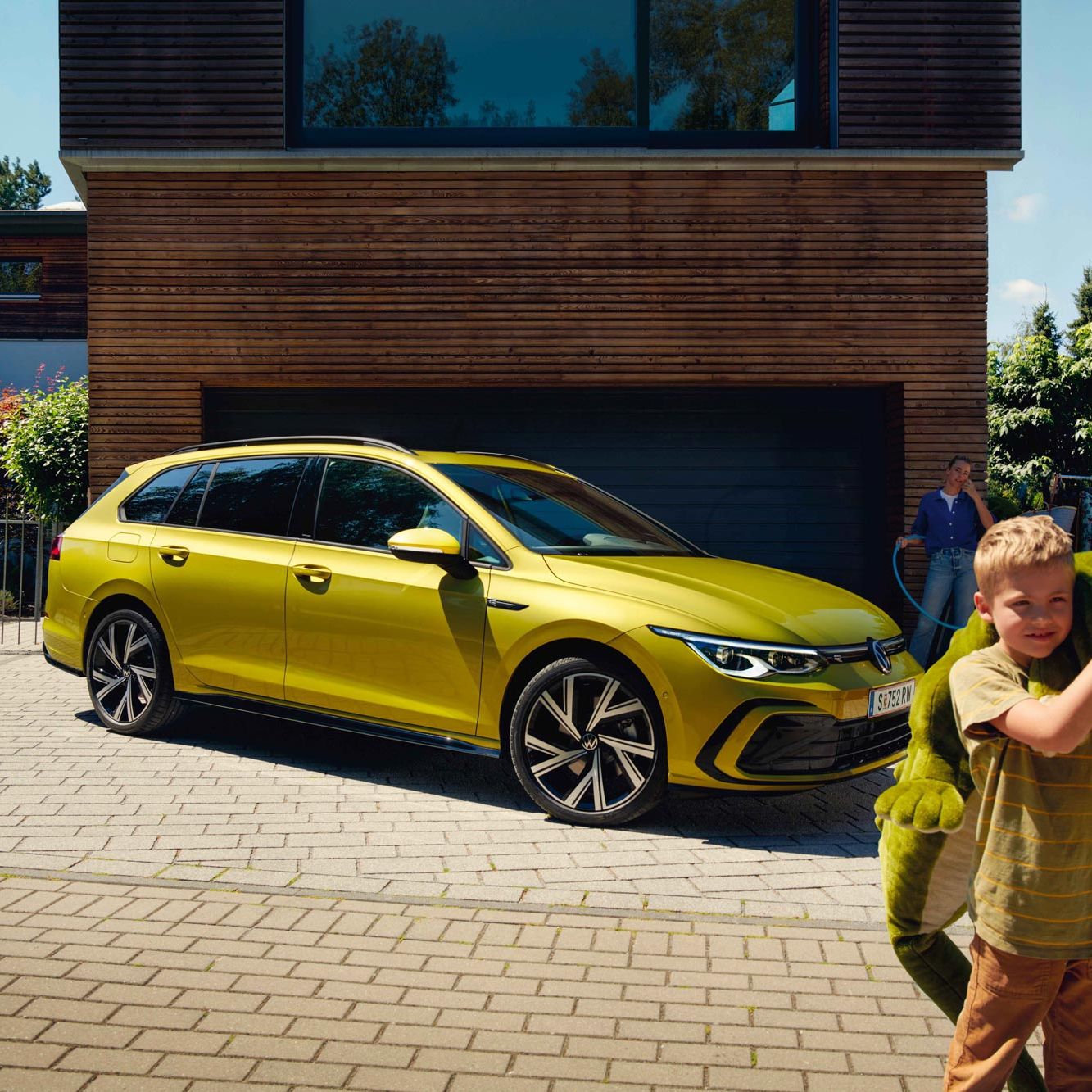 Момче с голяма плюшена играчка пред жълт Volkswagen Golf Variant, паркиран пред модерен дом