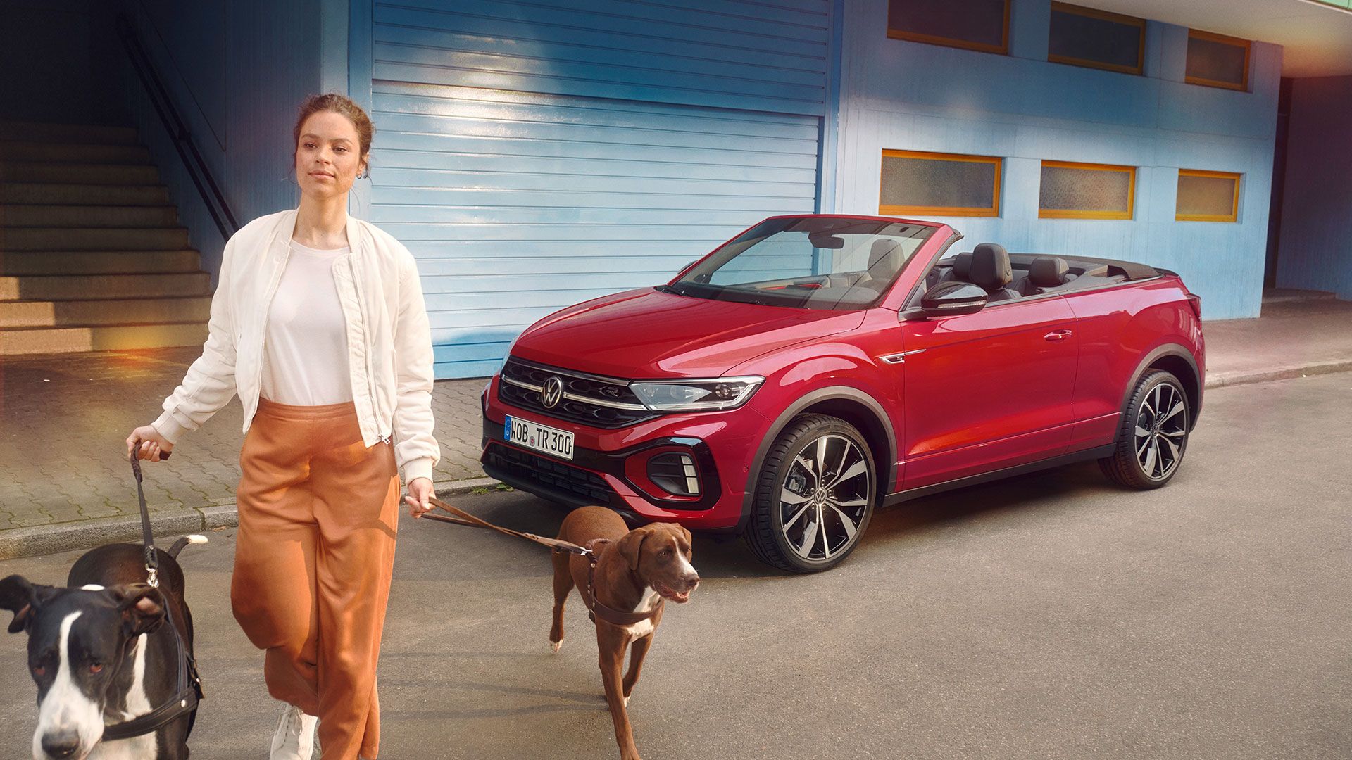 Ein rotes T-Roc Cabriolet parkt am Straßenrand mit geöffnetem Dach, eine Frau mit zwei Hunden überquert davor die Straße.