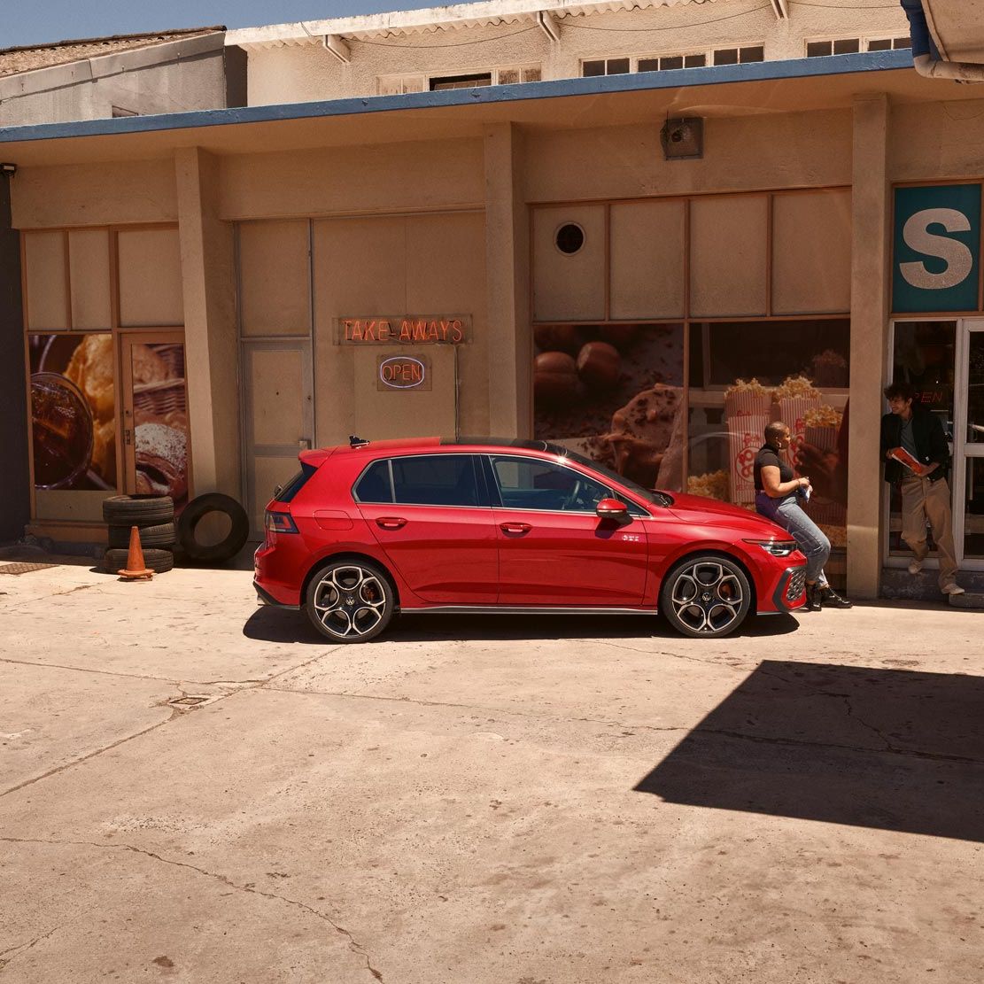 Червен Volkswagen Golf GTI паркиран пред малък магазин с хора, които разговарят на сянка