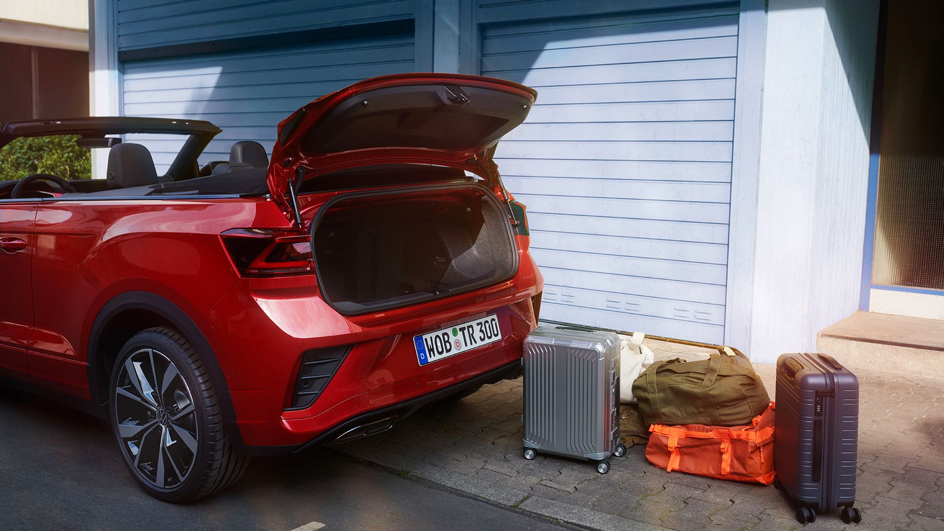 Ein VW T-Roc Cabriolet in rot parkt am Straßenrand. Blick seitlich auf den geöffneten Kofferraum, es stehen zwei Koffer und zwei Reisetaschen daneben. 