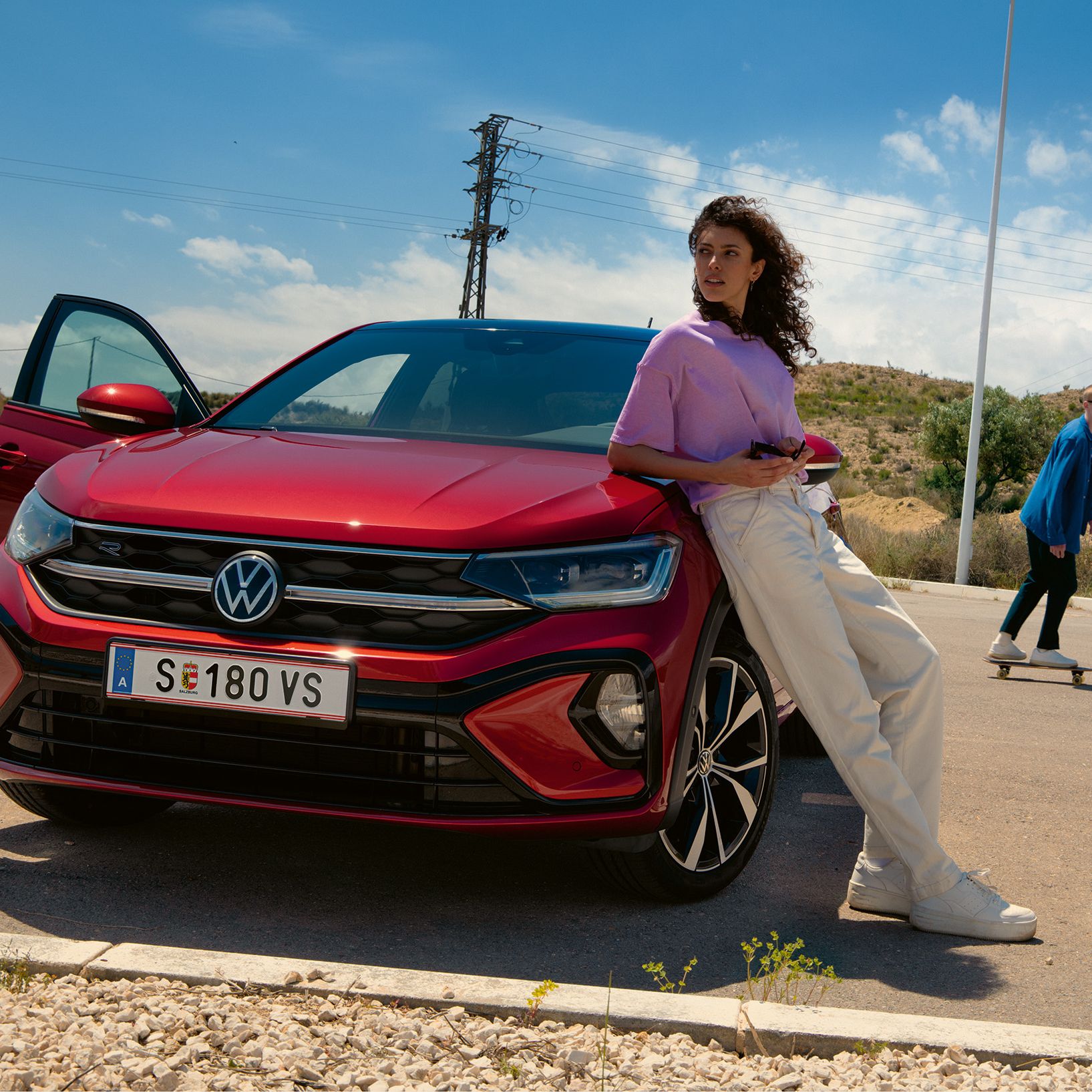 VW Taigo R-Line in Rot, Frontansicht auf Parkplatz. Frau lehnt an Motorhaube, Mann fährt Skateboard