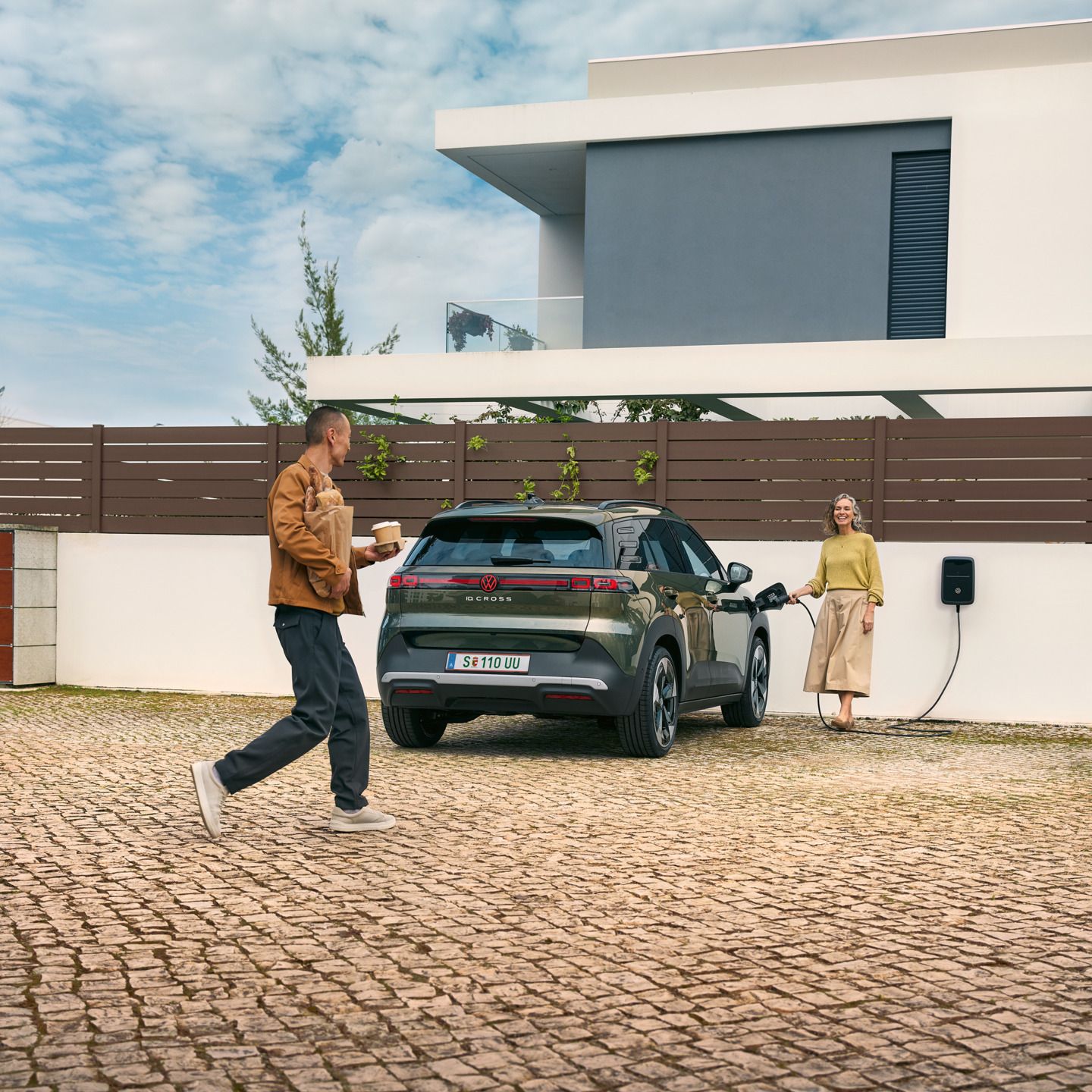 Жена включва кабел за зареждане от стенна кутия в зелен VW ID. Cross, мъж, се приближава до нея с чаши за кафе в ръка и хляб под мишница.
