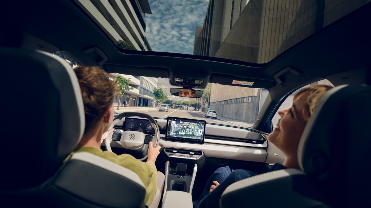 Мъж кара VW ID. Поло стил, на пасажерската седалка жена се смее. Високи сгради могат да се видят през панорамния покрив