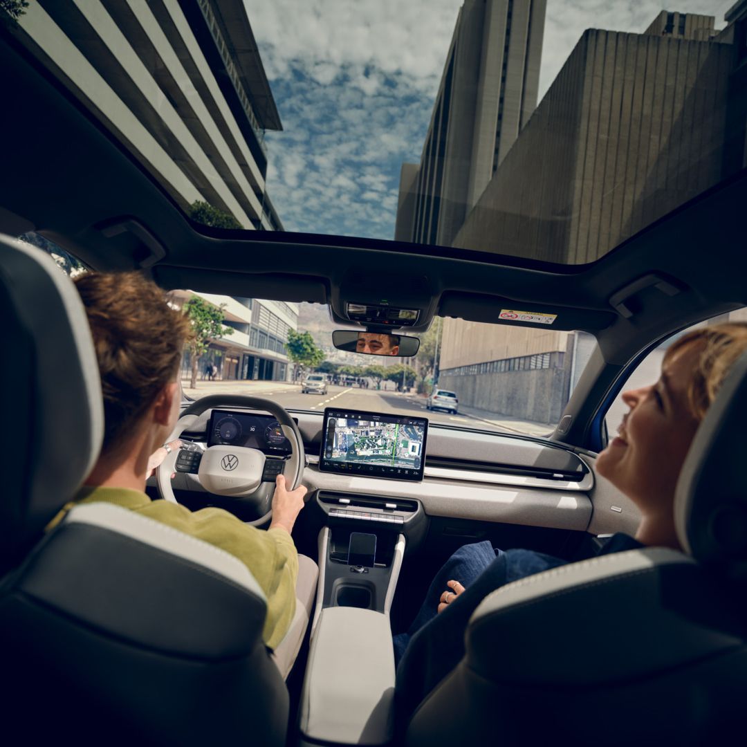 Мъж кара VW ID. Поло стил, на пасажерската седалка жена се смее. Високи сгради могат да се видят през панорамния покрив