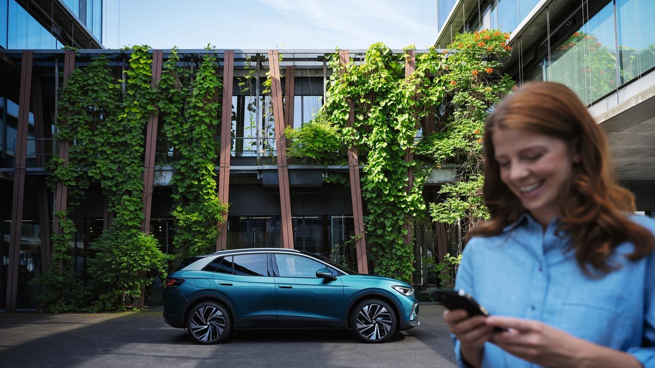 Junge Frau steht im Hof vor einem VW ID.4 Pro. Sie hält ein Handy in der Hand.
