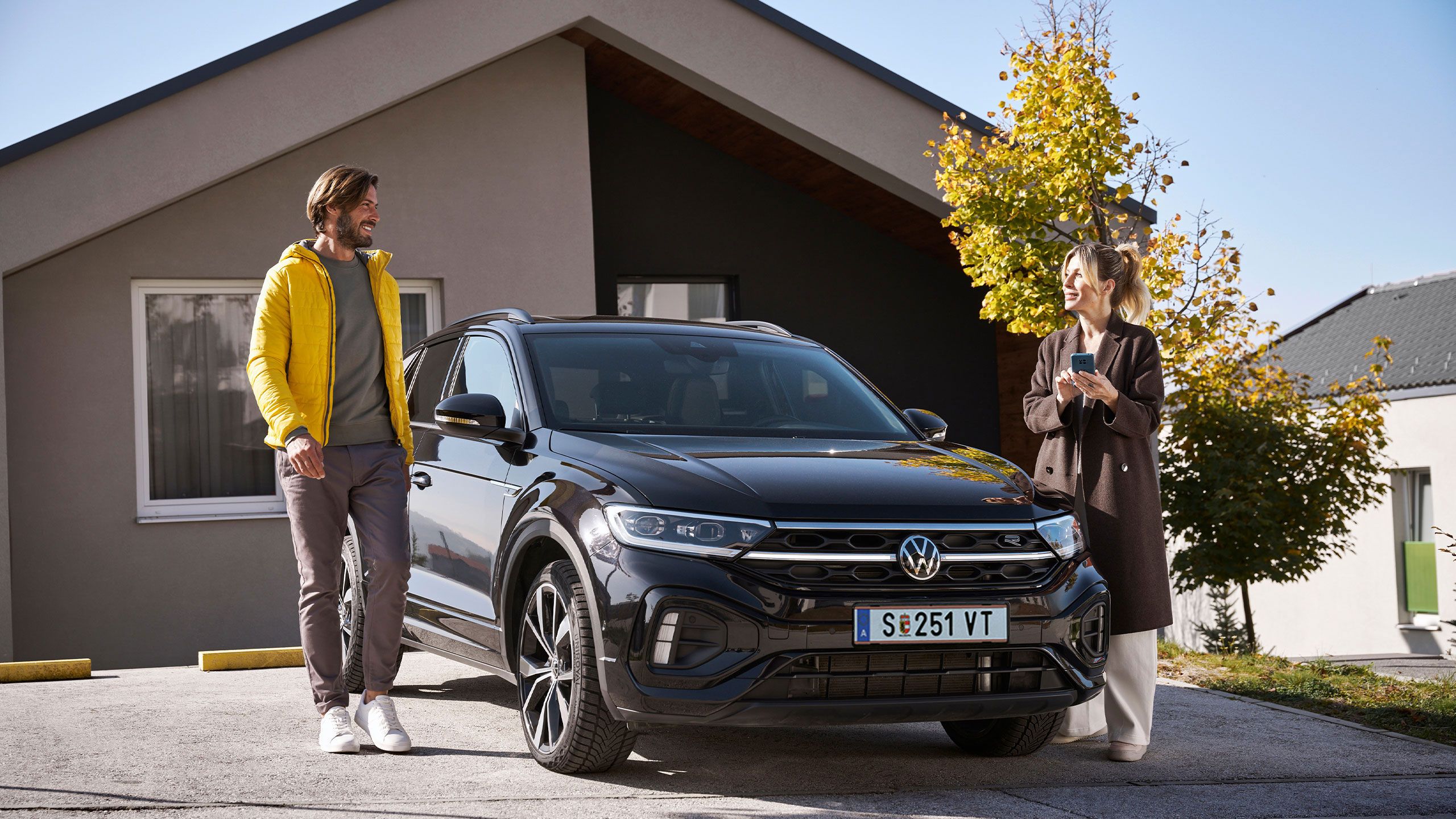 Мъж и жена стоят и разговарят до черен SUV Volkswagen, паркиран пред модерна къща.