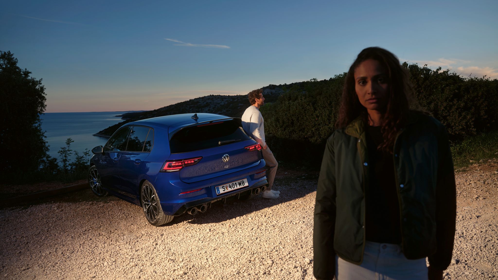 Frau steht im Vordergrund und dahinter ist ein Mann seitlich an den Golf R in Heckansicht angelehnt mit Blick auf Meer.