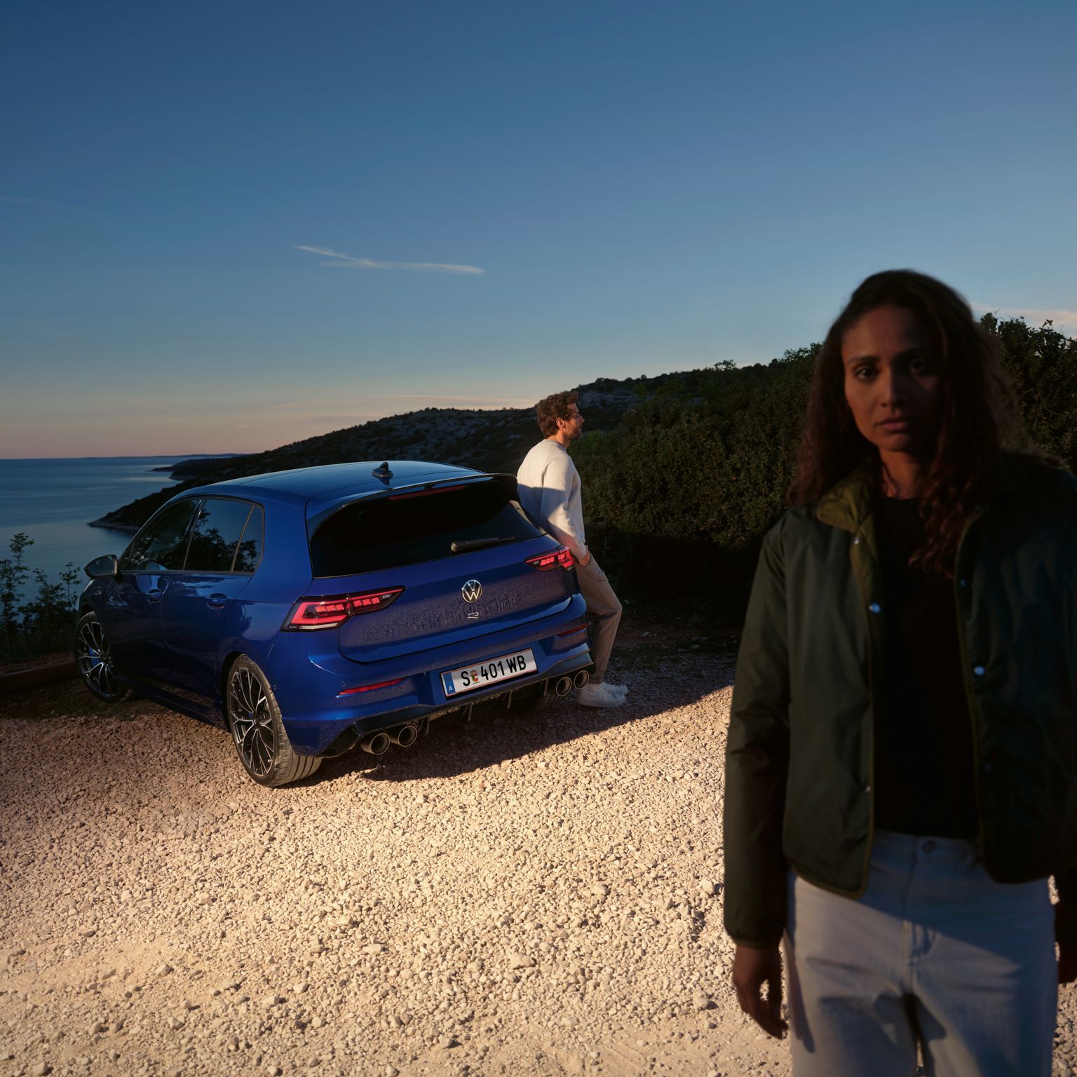 Frau steht im Vordergrund und dahinter ist ein Mann seitlich an den Golf R in Heckansicht angelehnt mit Blick auf Meer.